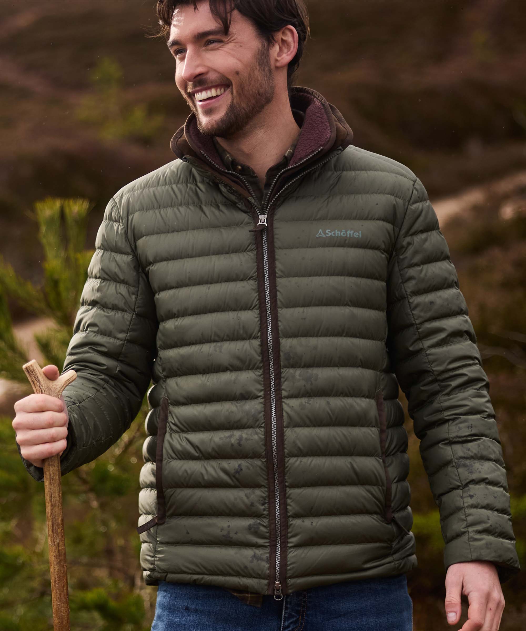 Man wearing olive green quilted down jacket with Schöffel logo, holding a wooden walking stick outdoors