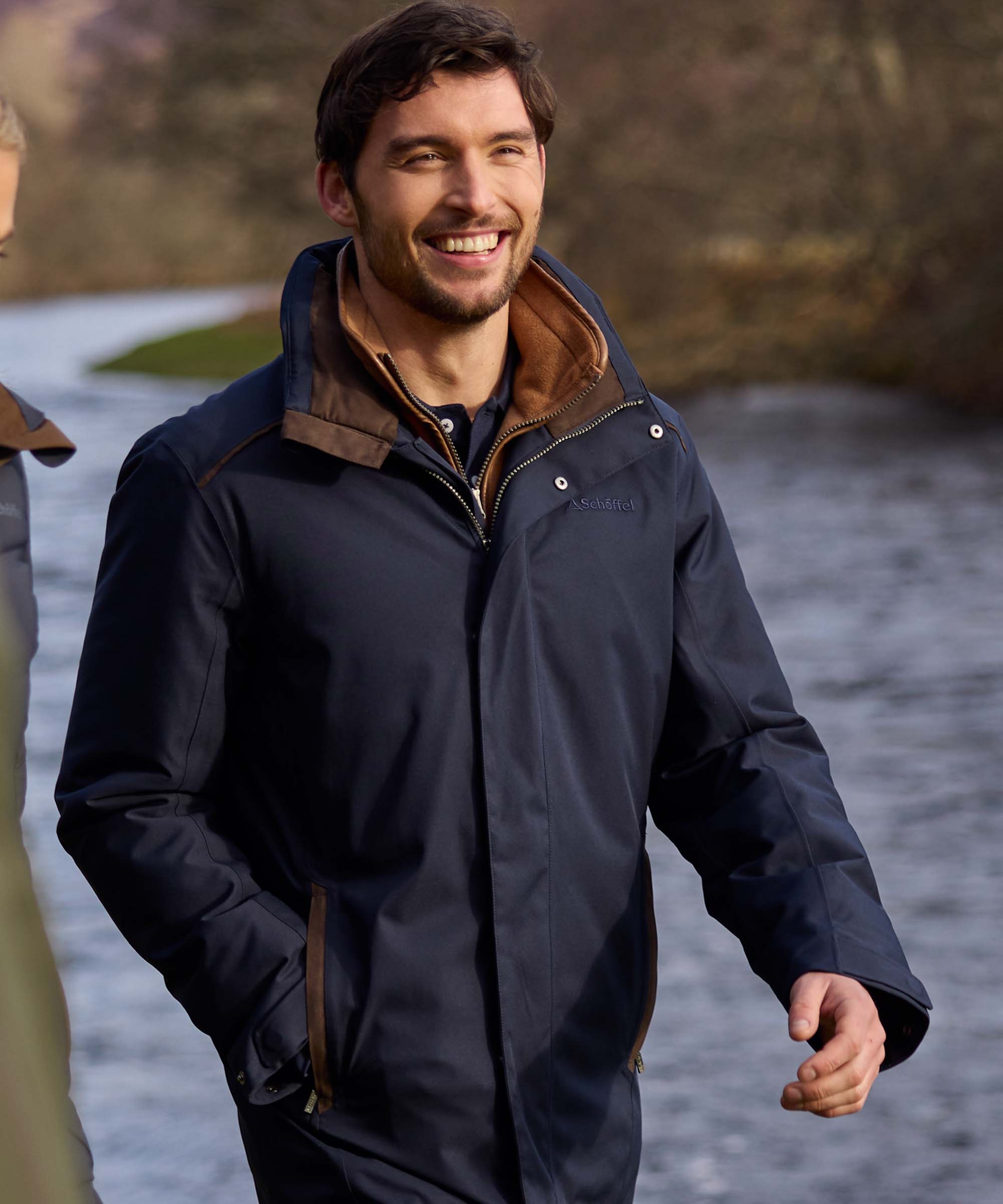 Man smiling in navy waterproof jacket with brown suede collar, walking beside a river
