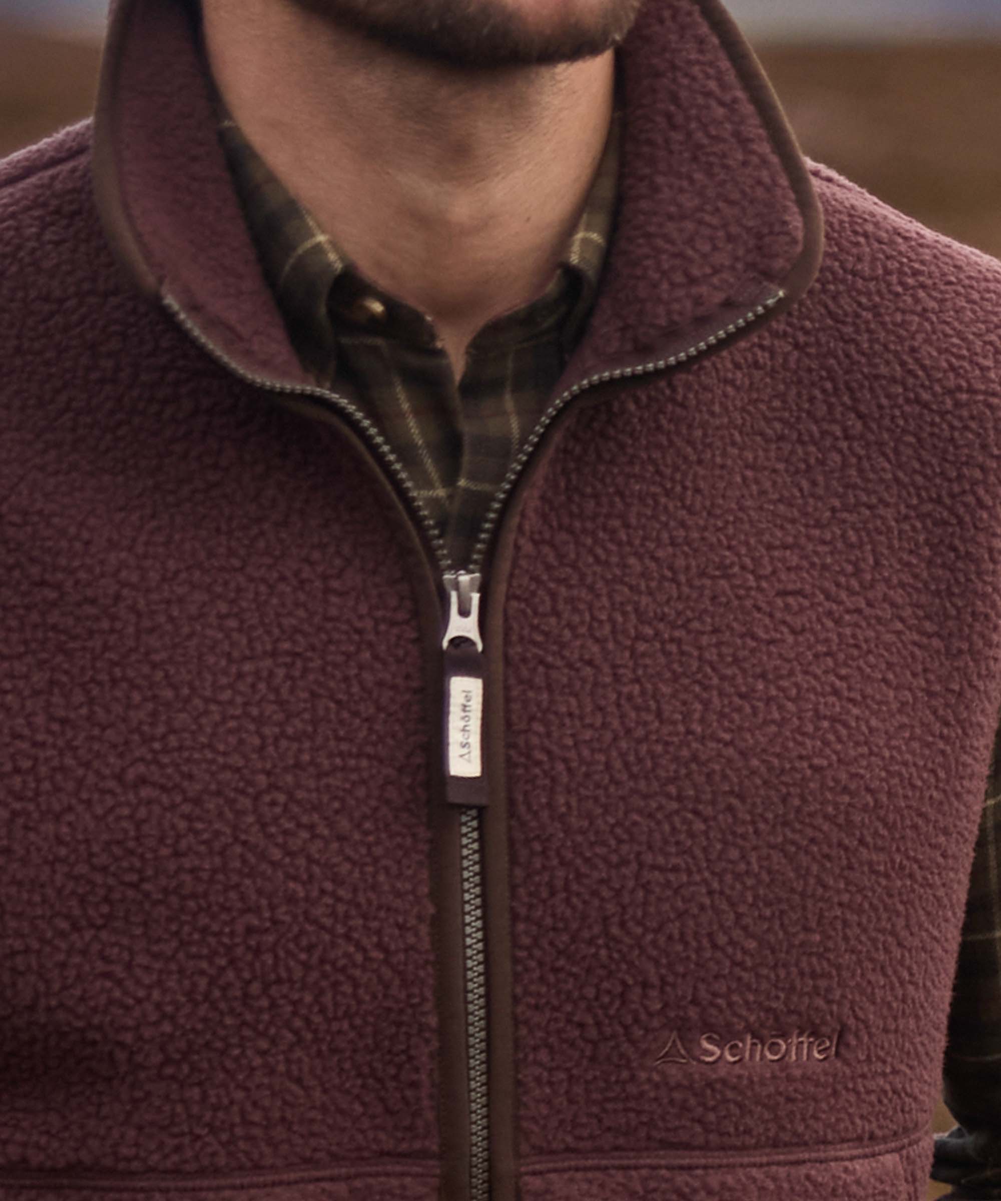 Close-up of burgundy sherpa fleece gilet collar and zip with Schöffel branding, worn over checked shirt