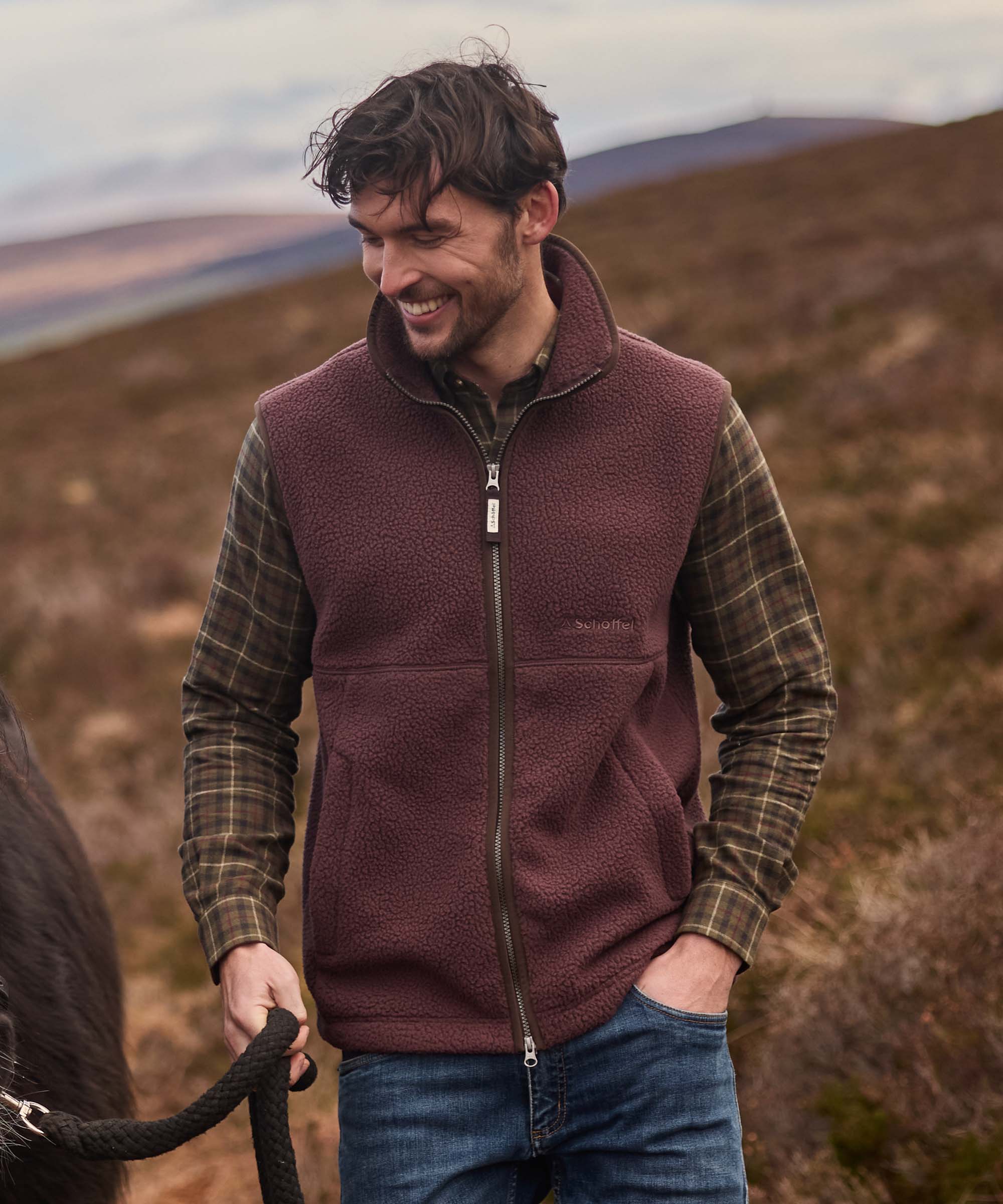 Man wearing burgundy sherpa fleece gilet over checked shirt, leading a horse across moorland
