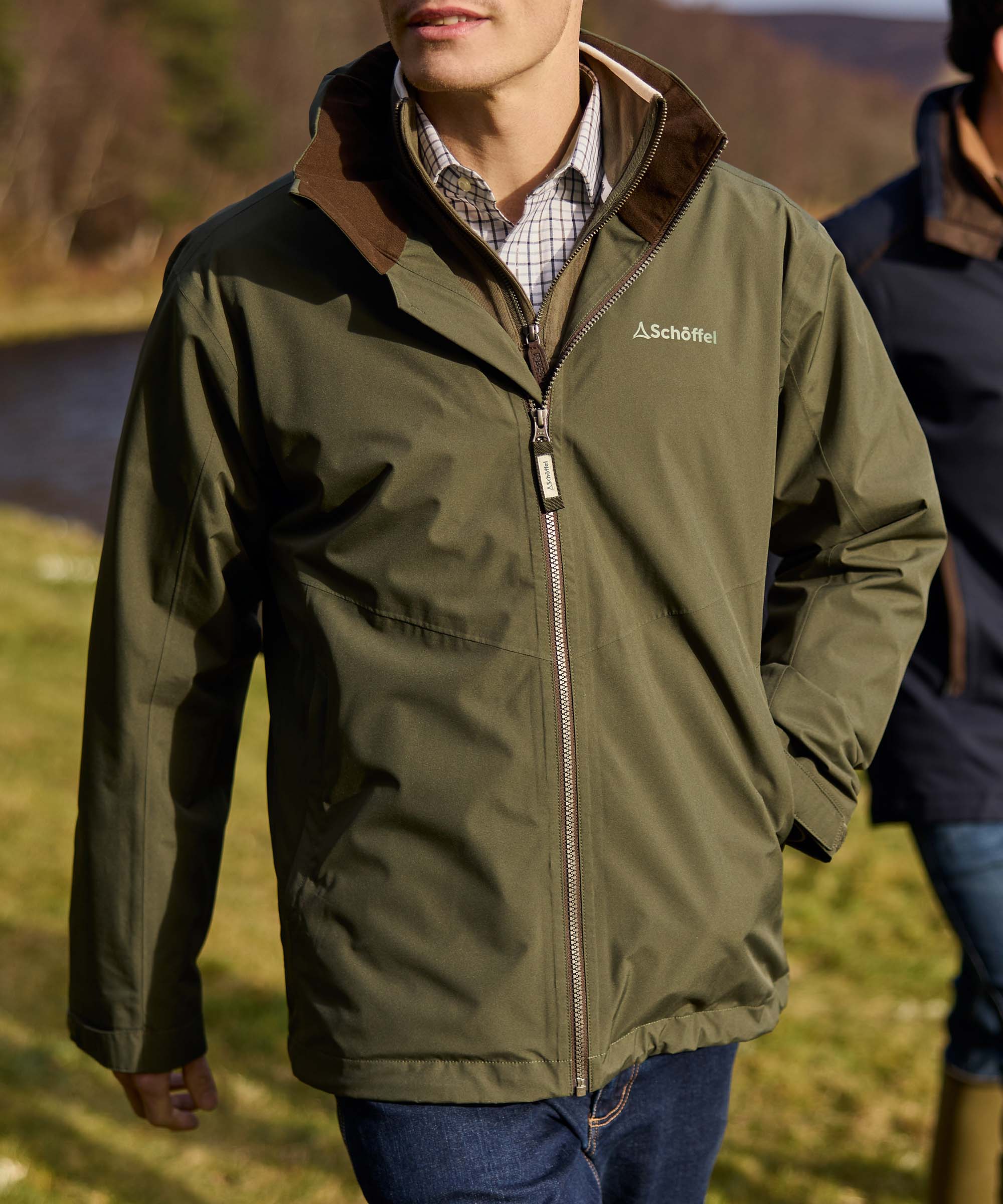 Man wearing olive green waterproof jacket with brown suede collar and Schöffel logo, countryside walk by riverside