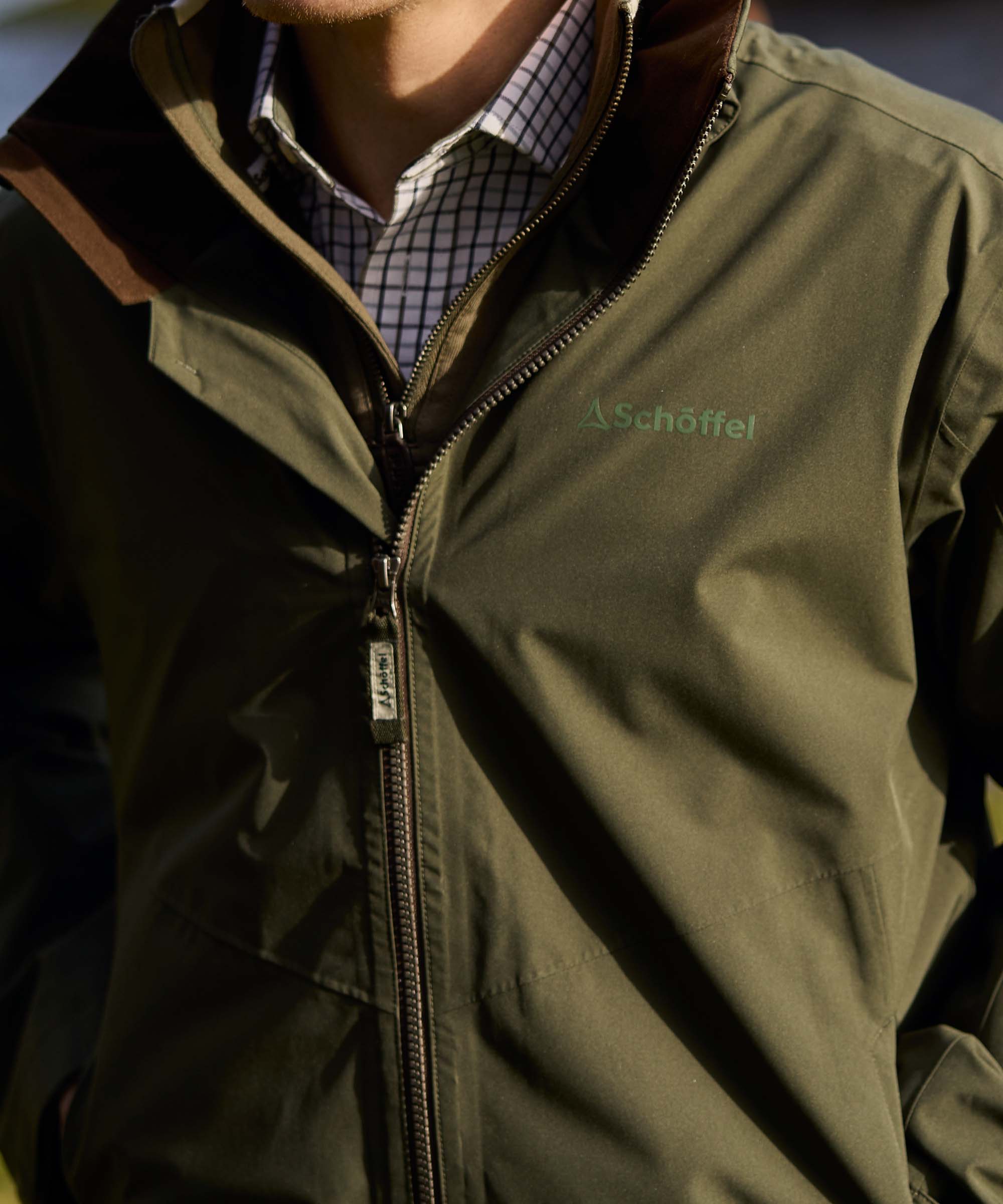 Detail of an olive green Schöffel jacket showing the Schöffel logo embroidered above the zip, worn with a checked shirt for country style.