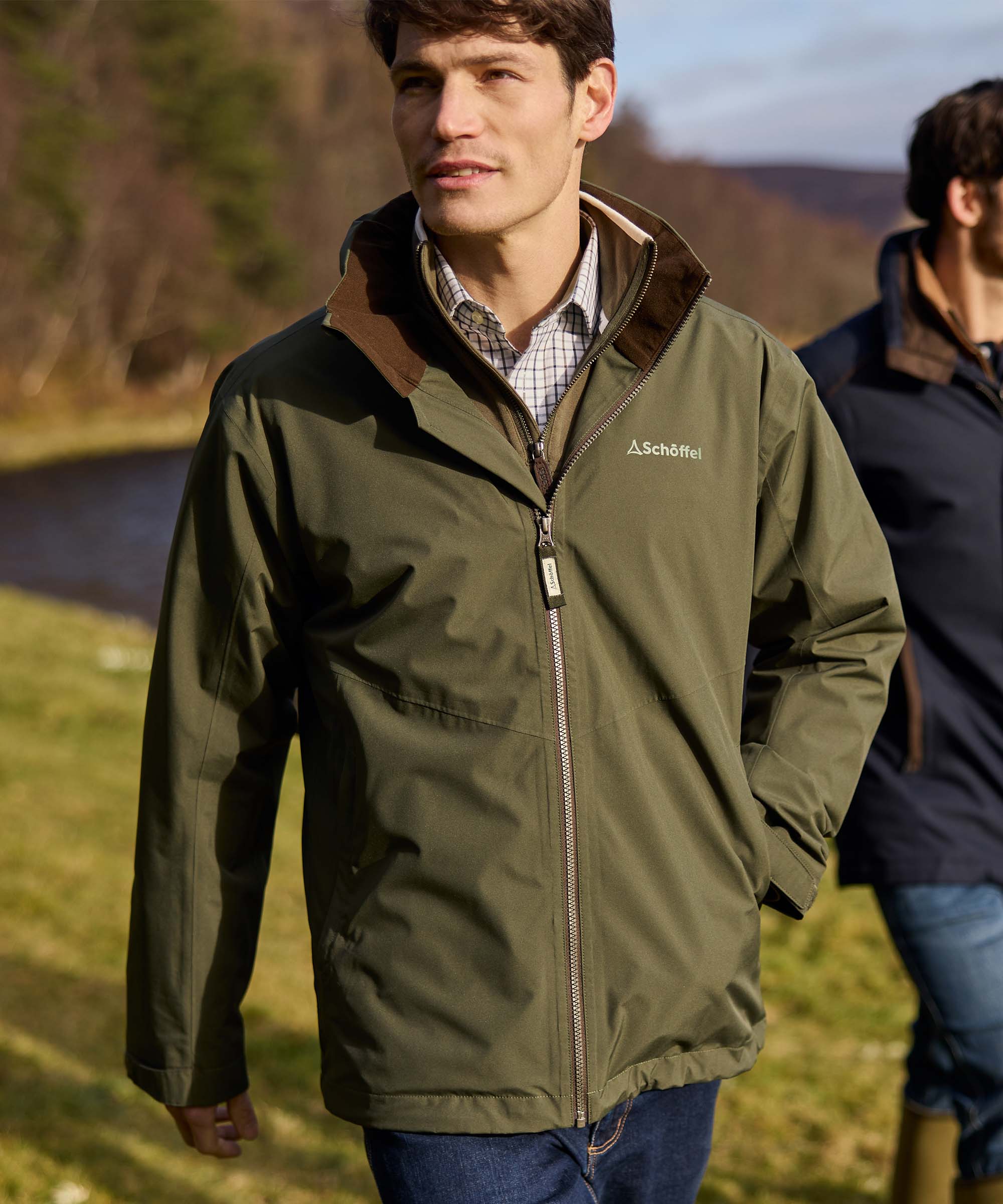 Man walking beside a river wearing an olive green Schöffel waterproof jacket with brown corduroy collar, checked shirt and jeans, styled for a countryside walk.