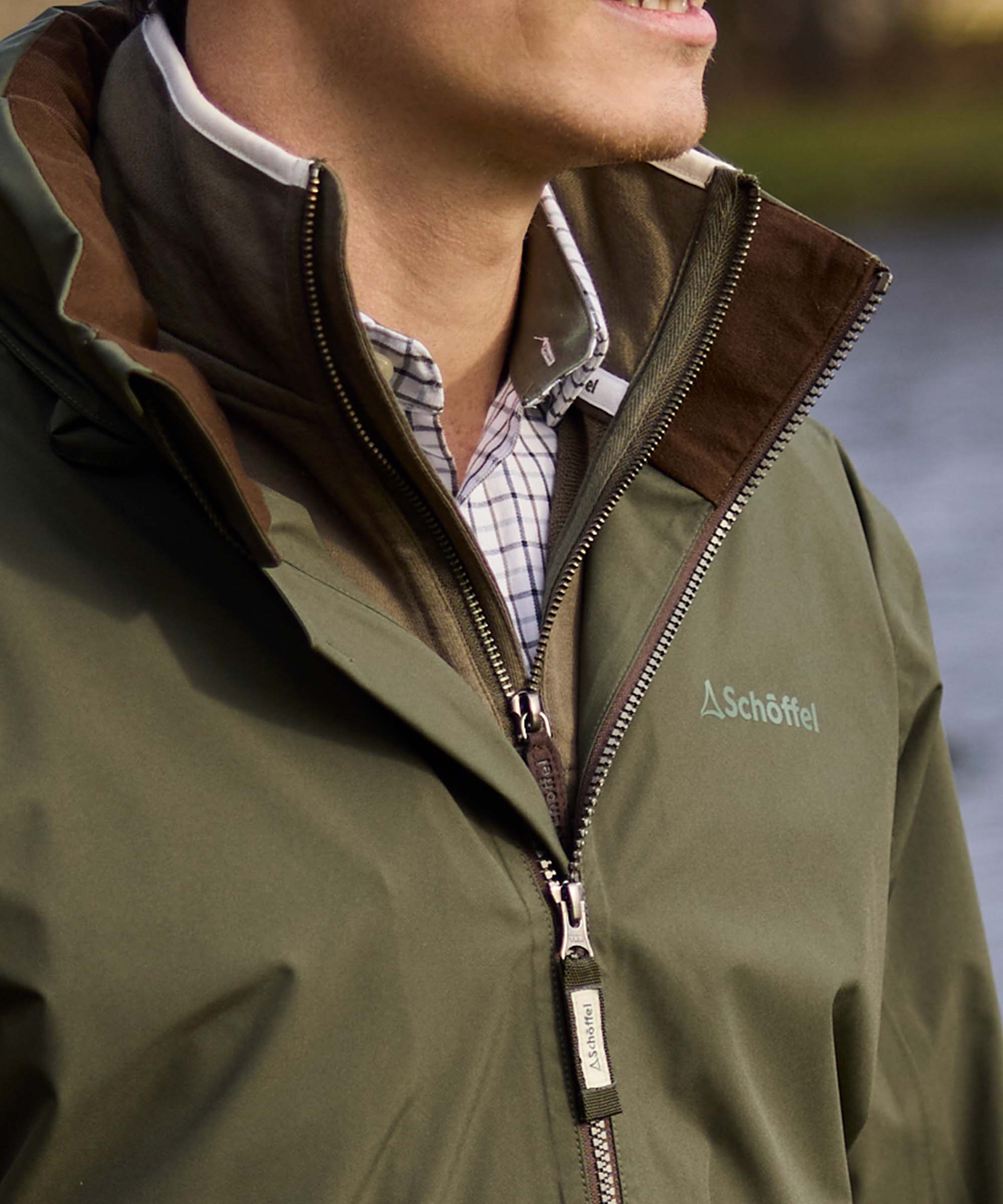 Close-up of an olive green Schöffel jacket collar with brown corduroy trim and branded zip pull, worn over a checked shirt for classic country style.
