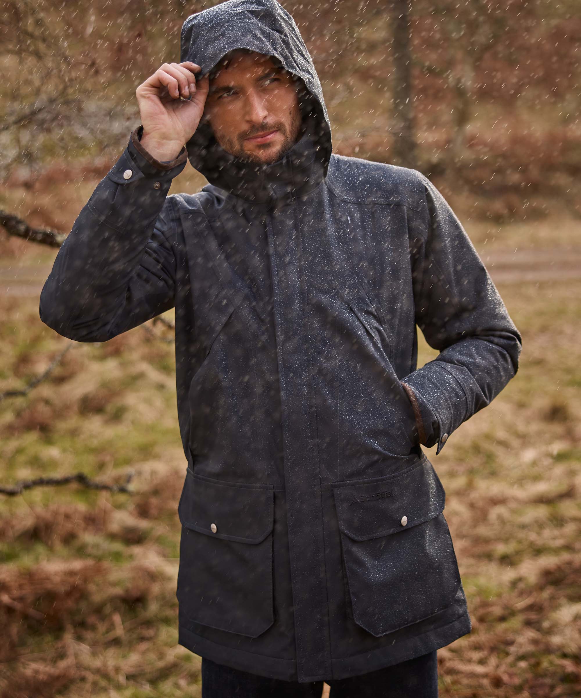 Man in a navy Schöffel waterproof jacket caught in the rain, pulling up the hood in a woodland setting.