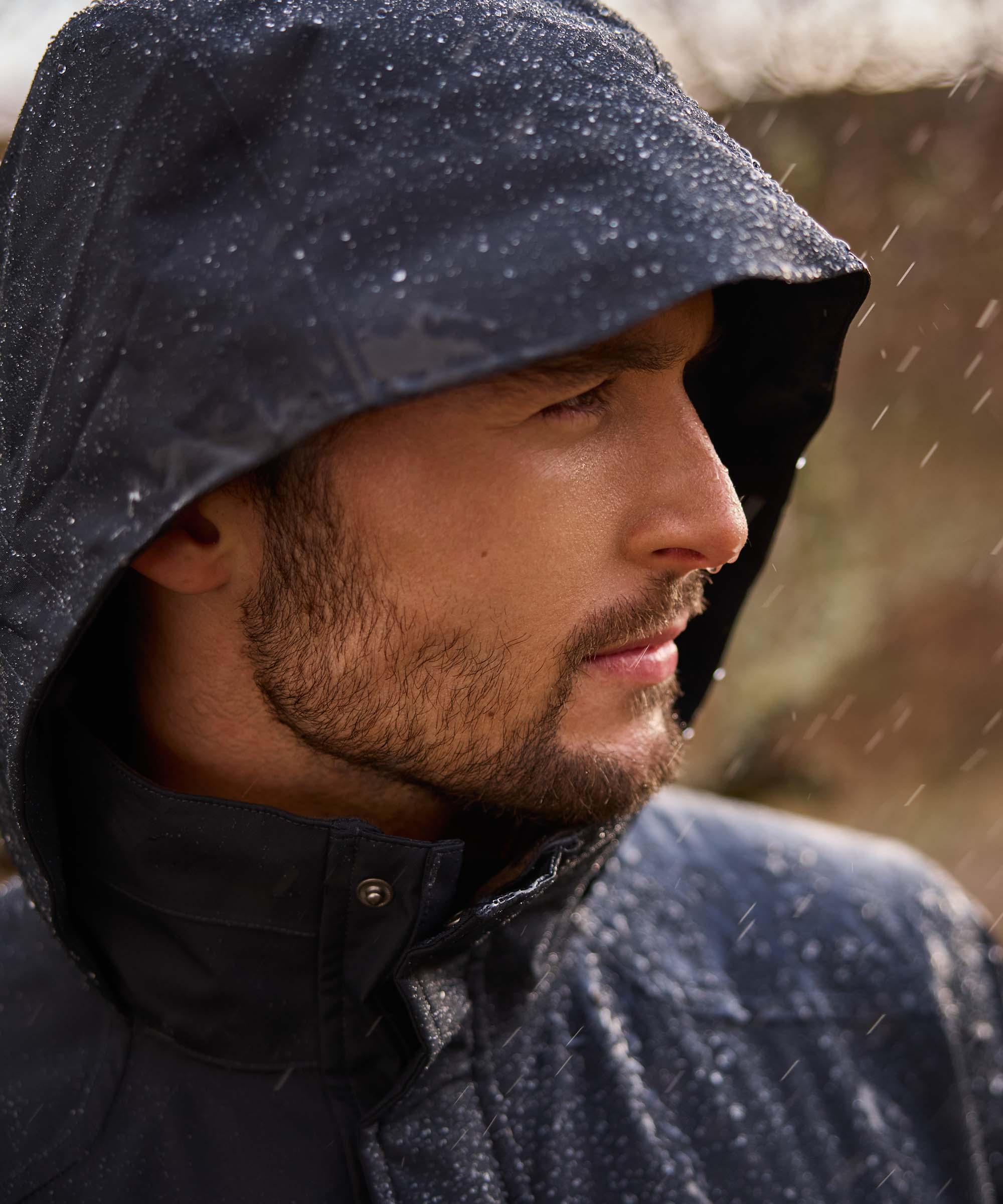 Close-up of a man in a navy Schöffel jacket with the hood up, sheltering from the rain with the collar fastened.