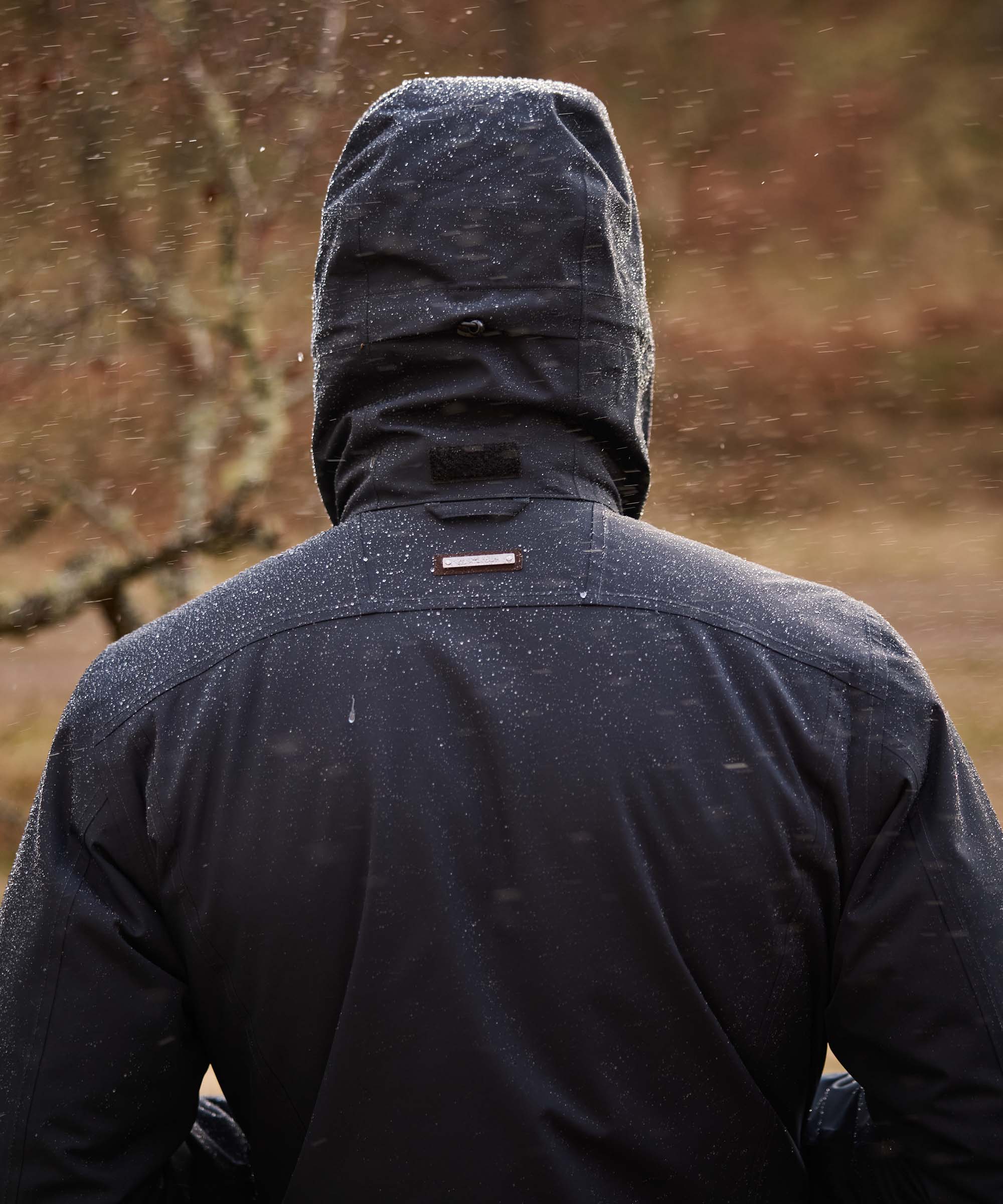 Rear view of a man in a navy Schöffel waterproof jacket with the hood up, walking through rainy woodland.