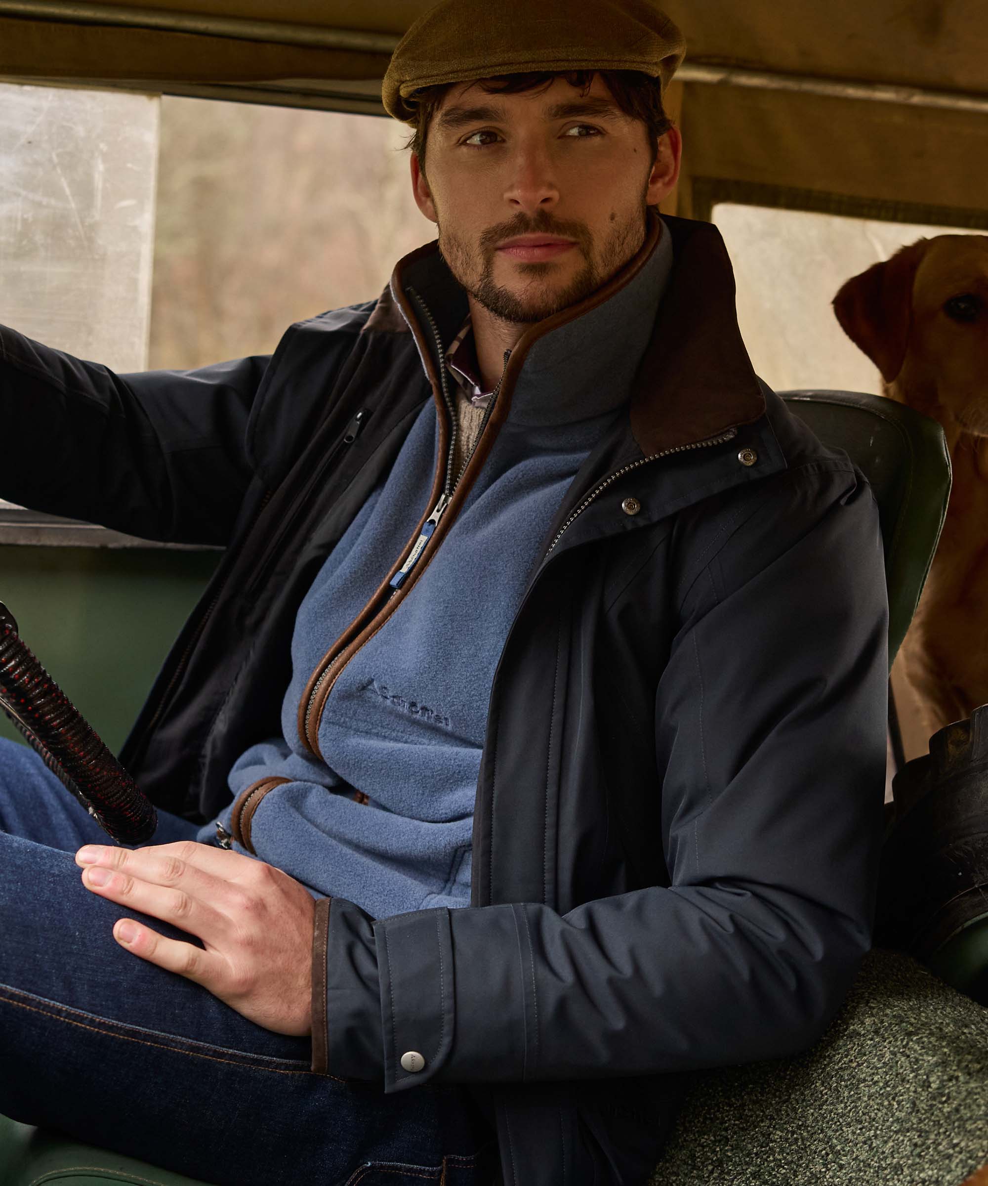Man sat in a Land Rover wearing a navy Schöffel waterproof jacket with brown corduroy collar, layered over a blue quarter-zip fleece, with a Labrador beside him.