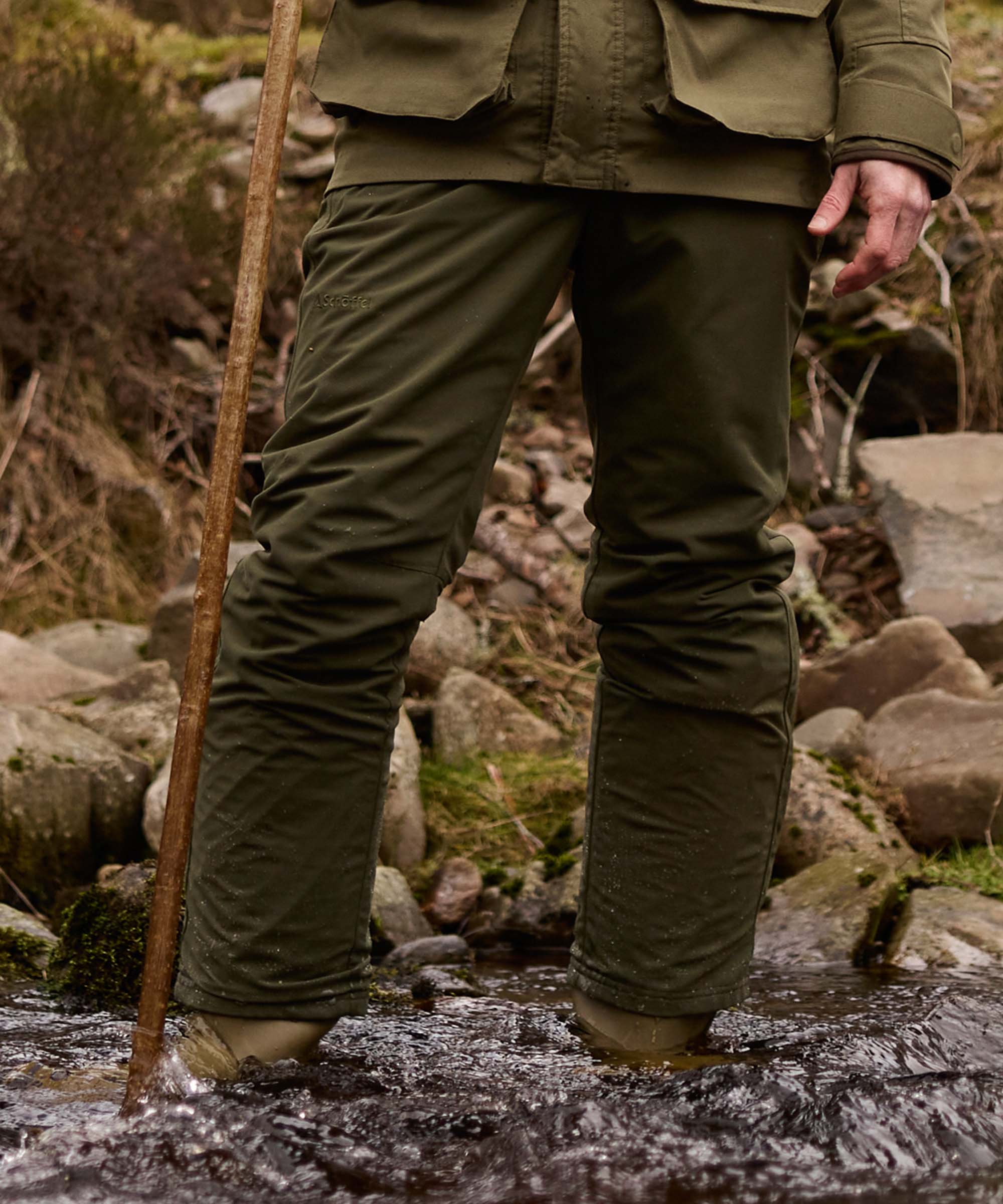 Rear view of a man's legs in olive green Schöffel waterproof trousers, wading through a rocky stream with a walking stick.