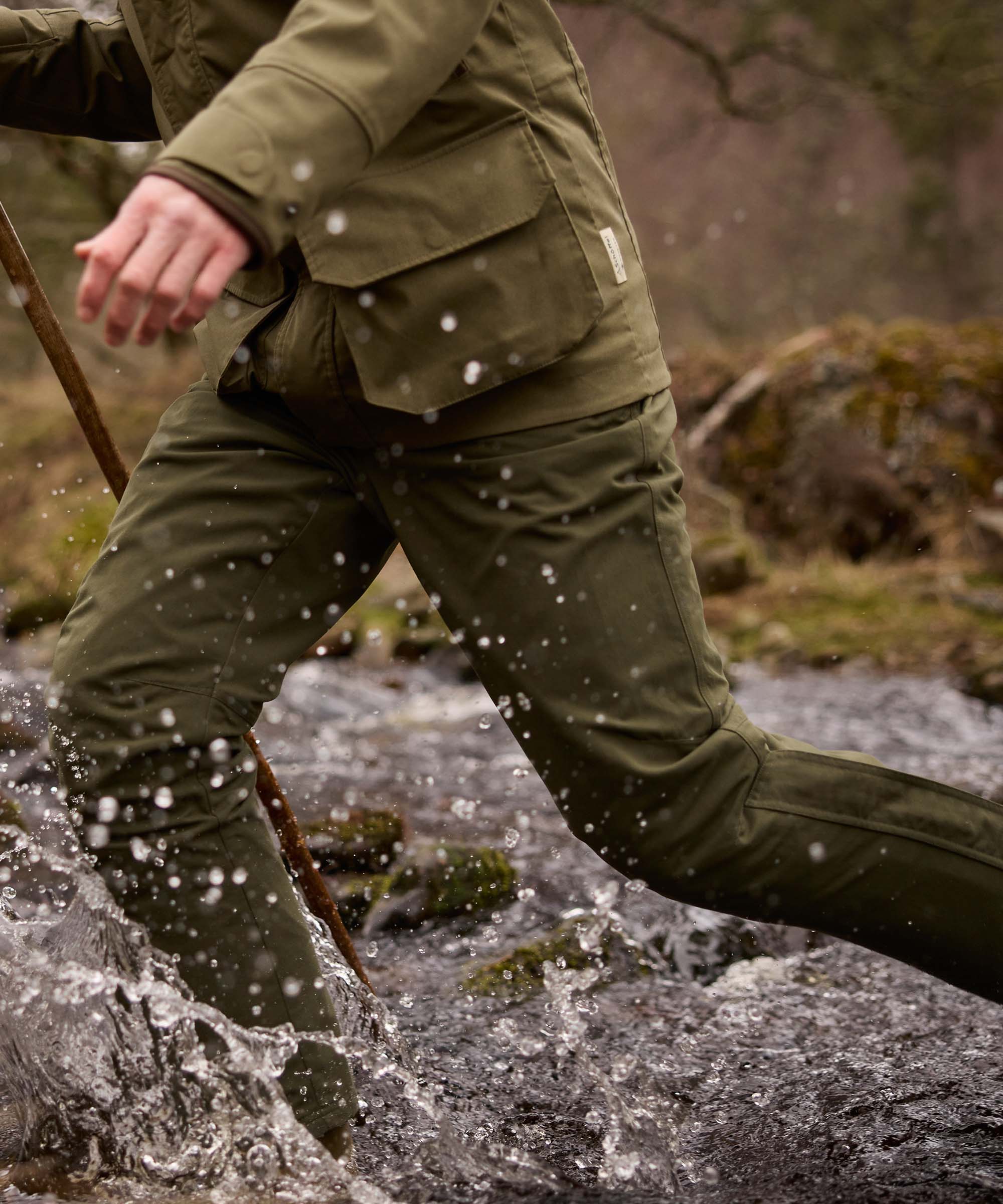 Man striding through a stream wearing an olive green Schöffel jacket and matching waterproof trousers, splashing through water with a walking stick.