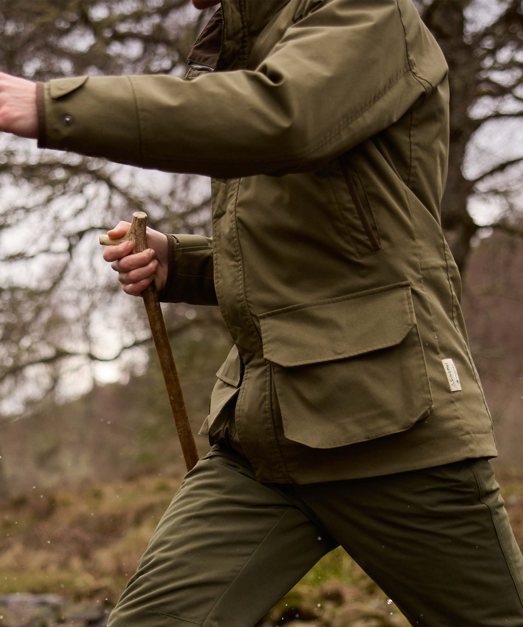 Man walking through woodland wearing an olive green Schöffel jacket, holding an antler-handled walking stick.