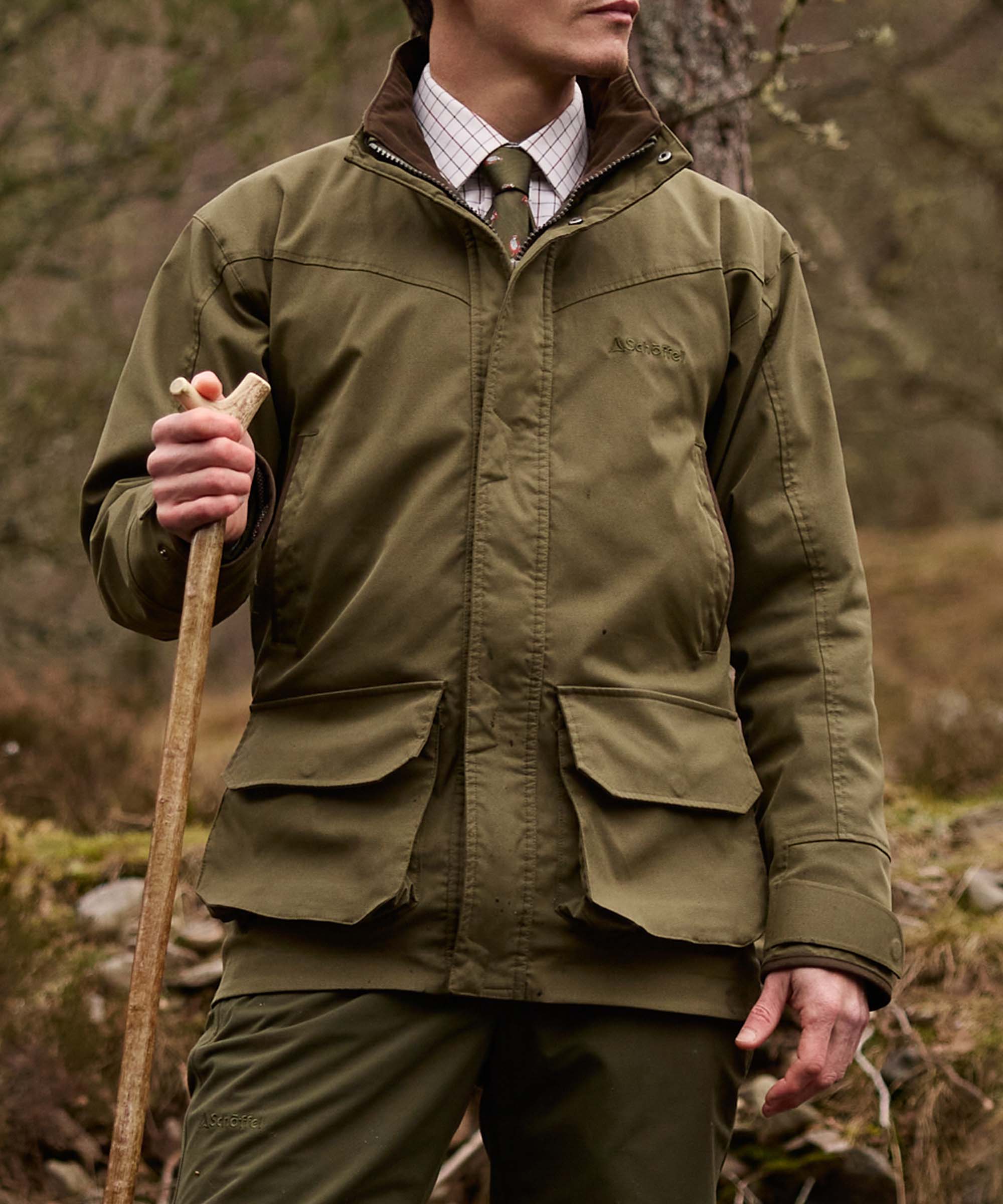 Man wearing an olive green Schöffel jacket and tie, holding an antler-handled walking stick in a woodland setting.