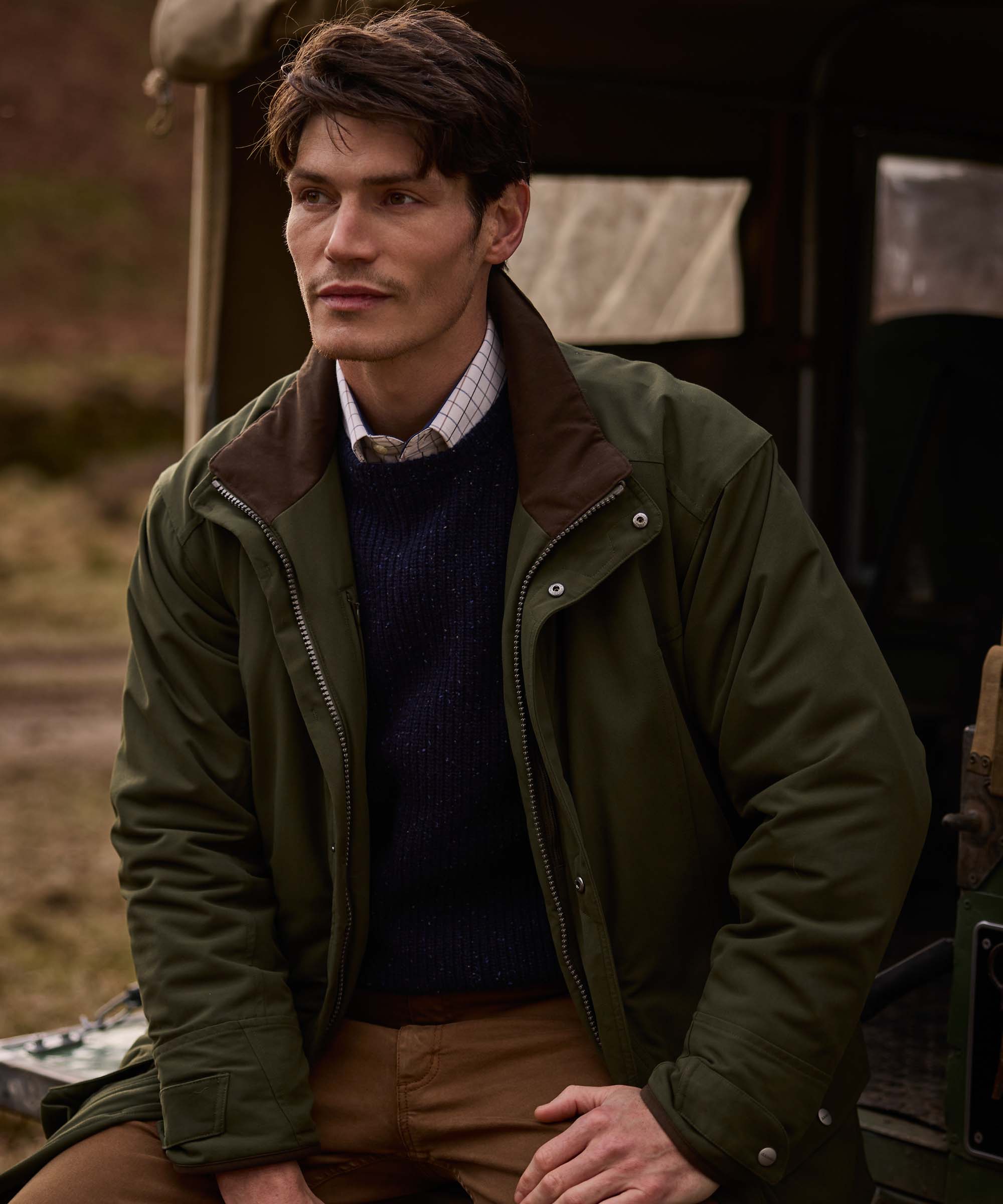 Man wearing an olive green Schöffel field jacket with contrast brown corduroy collar, layered over a navy jumper and checked shirt, styled with moleskin trousers beside a Land Rover in the Scottish countryside.