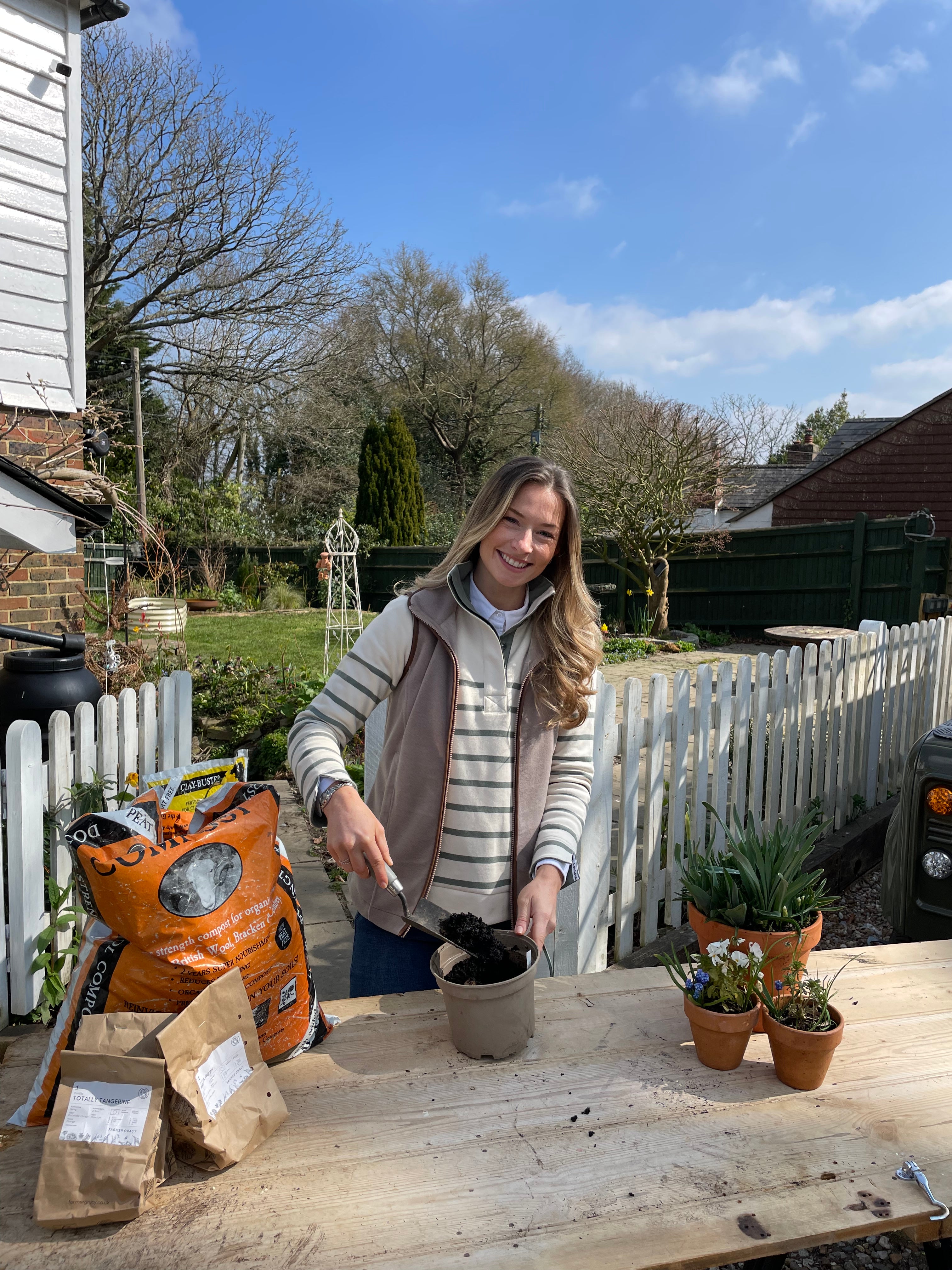 Person standing outdoors with gardening tools and plants, under a clear blue sky.