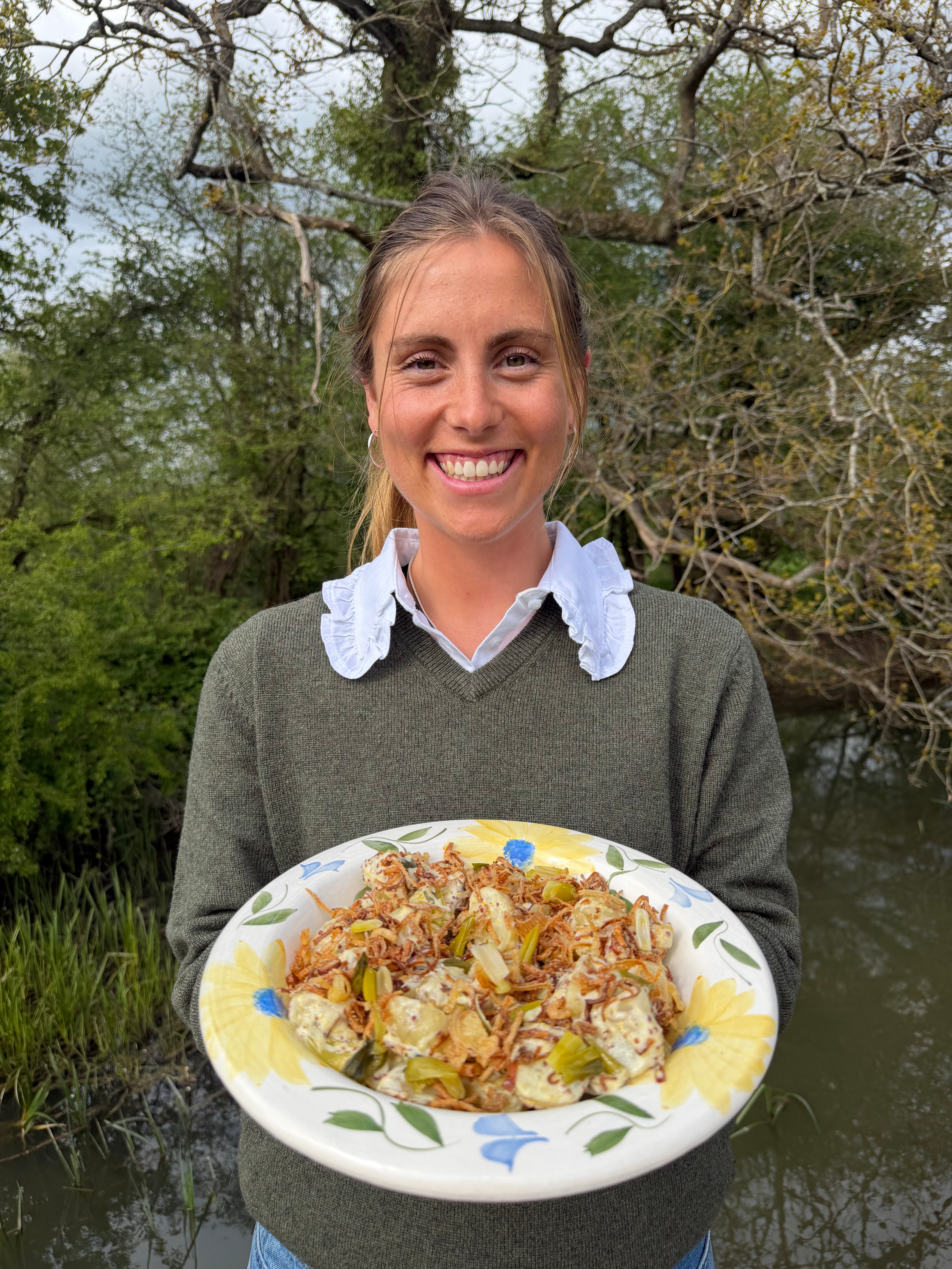 Person holding a plate of food outdoors with trees and water in the background