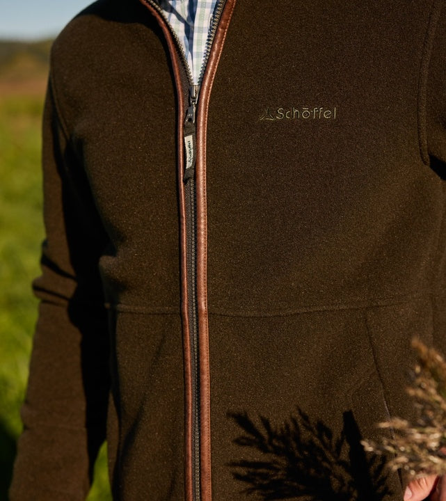 Close-up of a man wearing a dark olive fleece quarter-zip pullover with tan leather-trim zip and embroidered Schöffel logo on the chest, layered over a checked shirt in an outdoor countryside setting.