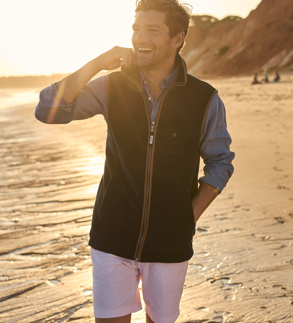 Man standing on a beach wearing a navy Oakham gilet and pink shorts, with a warm glow indicating sunset.