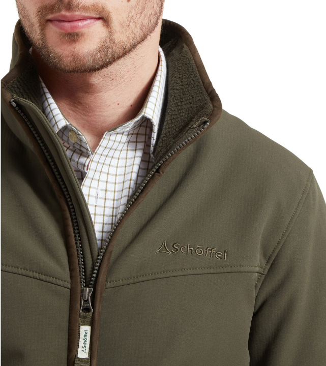 Close-up of a man wearing a forest green softshell jacket with a fleece-lined collar, brown suede-effect trim, and a tonal embroidered Schöffel logo on the chest. A branded zip pull is visible at the hem of the zip. A green and white windowpane check shirt is visible underneath.