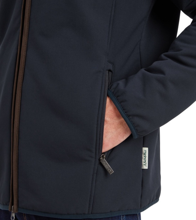 Close-up detail of a man's hand in the zipped side pocket of a navy softshell jacket, showing the tan leather-trim zip and branded Schöffel zip pull, worn with blue jeans.