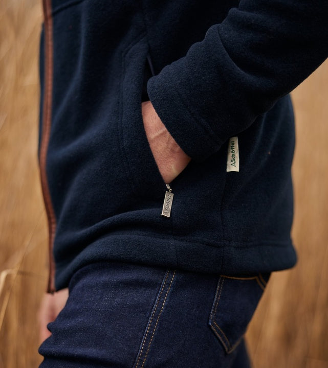 Close-up detail of a man's hand in the zipped side pocket of a navy fleece jacket with tan leather-trim zip and branded Schöffel zip pullers, worn with dark blue jeans.