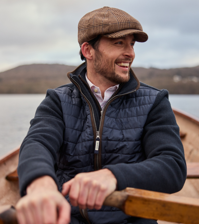 Man wearing a navy quilted jacket and plaid cap, sitting in a boat with mountains in the background.