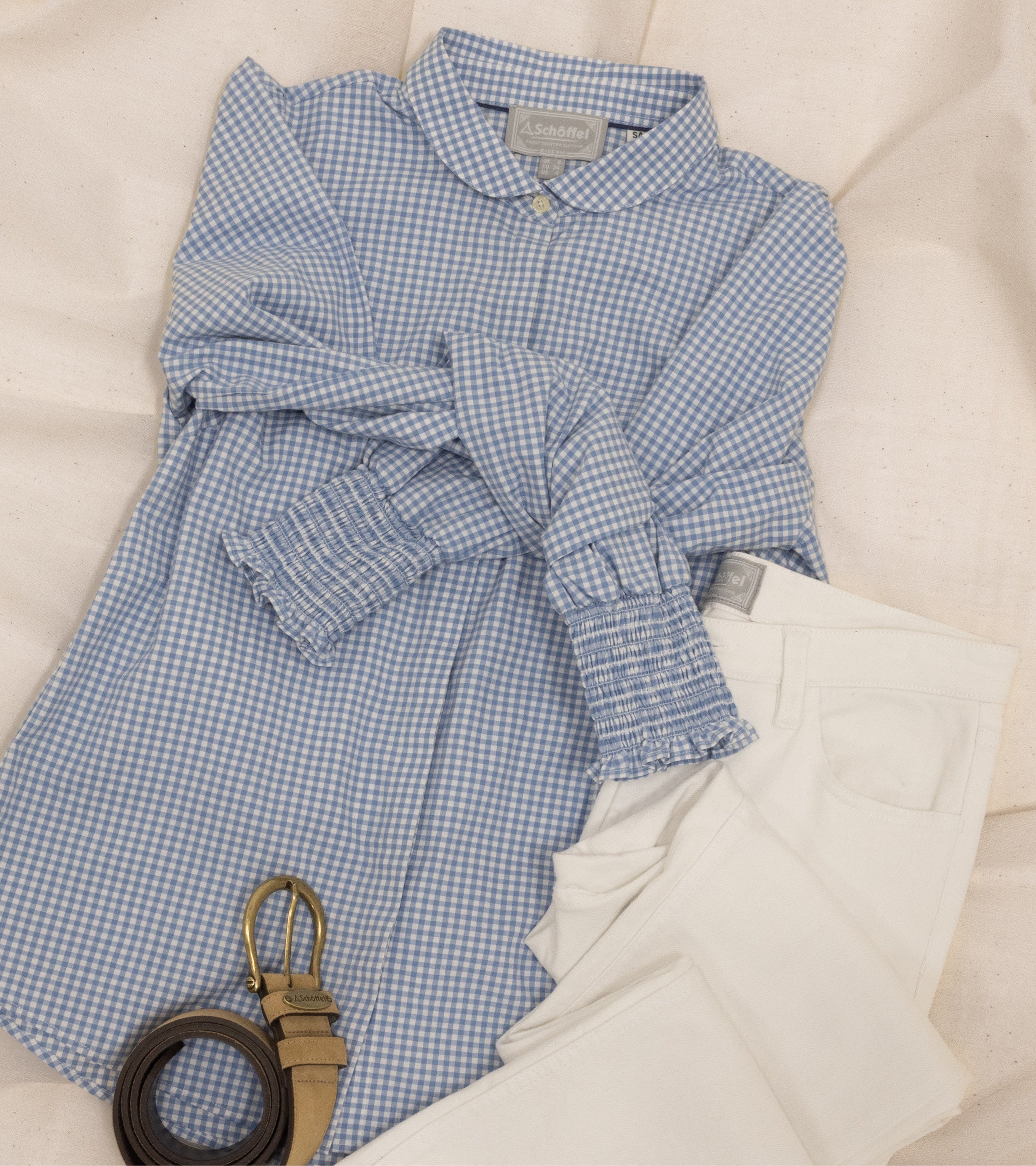 A blue and white gingham blouse with a Peter Pan collar and smocked cuffs is neatly laid out on a soft, neutral fabric background. The sleeves are gently tied together at the front. Alongside the blouse, a pair of off-white trousers with a visible brand label is folded. A brown leather belt with a gold buckle is coiled next to the outfit, completing the laid-back yet sophisticated look.