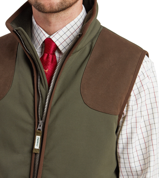 Close-up of the upper front of a forest green field jacket with a zip fastening, a fleece-lined collar, and tan suede shoulder panel. A white windowpane check shirt and an olive herringbone tie with a pheasant print are visible underneath. A Schöffel branded zip pull is shown at the chest.