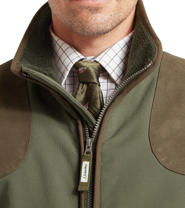 Close-up of the upper front of a forest green field jacket with a zip fastening, a fleece-lined collar, and tan suede shoulder panel. A white windowpane check shirt and an olive herringbone tie with a pheasant print are visible underneath. A Schöffel branded zip pull is shown at the chest.