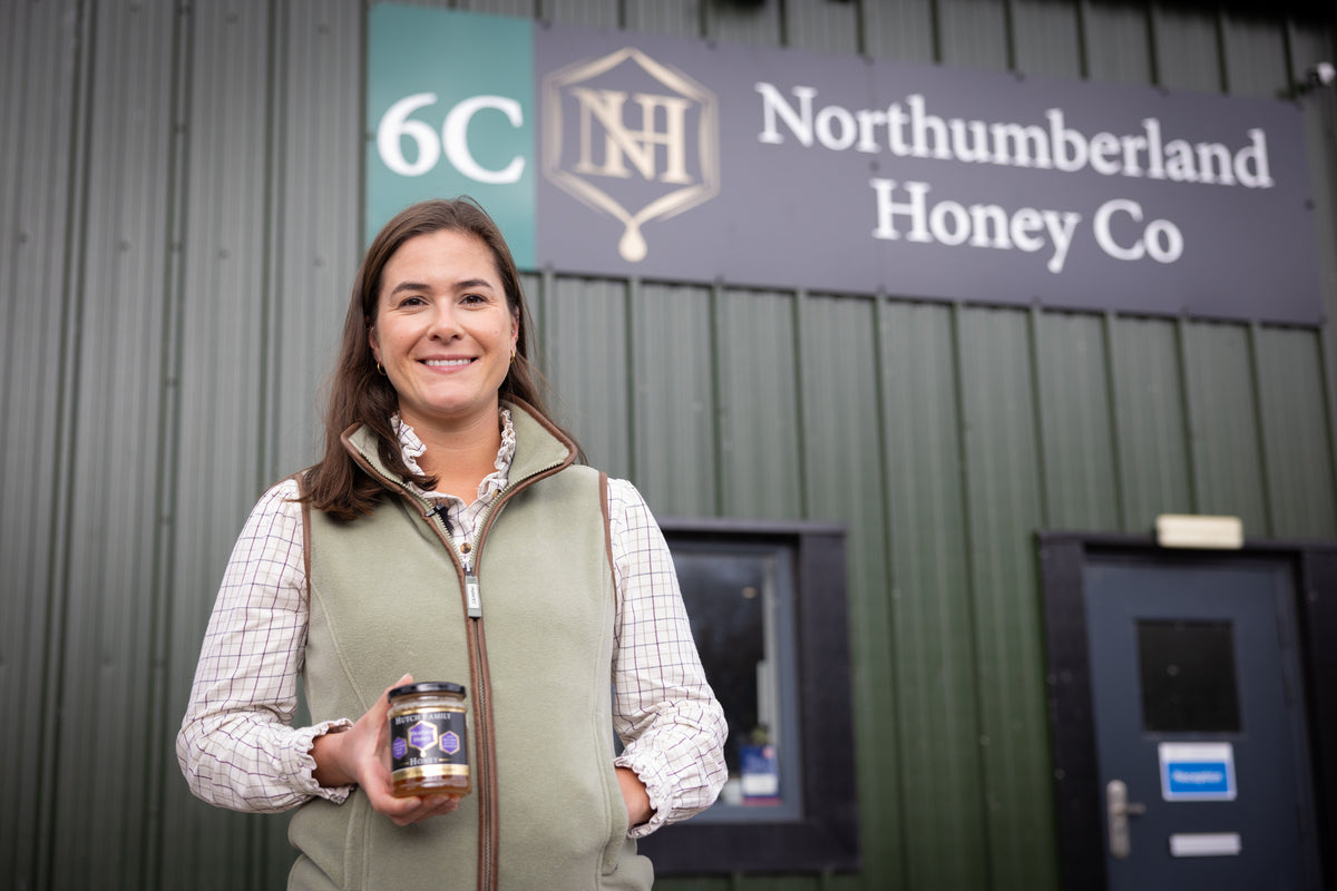 A woman standing in front of green building holding a jar of British honey.