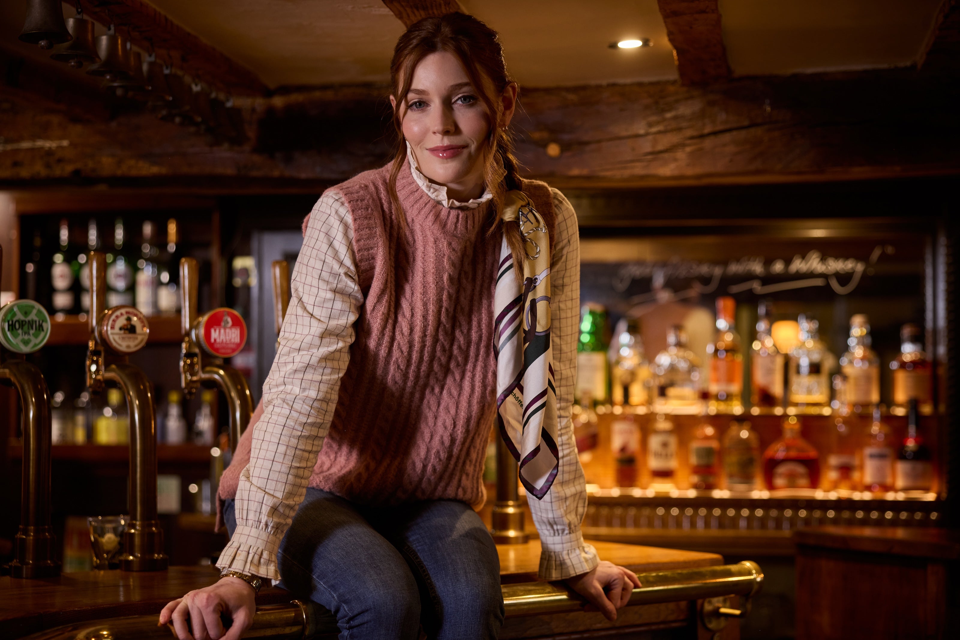 A woman wearing a pink cable-knit sleeveless vest over a beige plaid shirt and a striped necktie sits on the counter of a warmly lit pub bar.