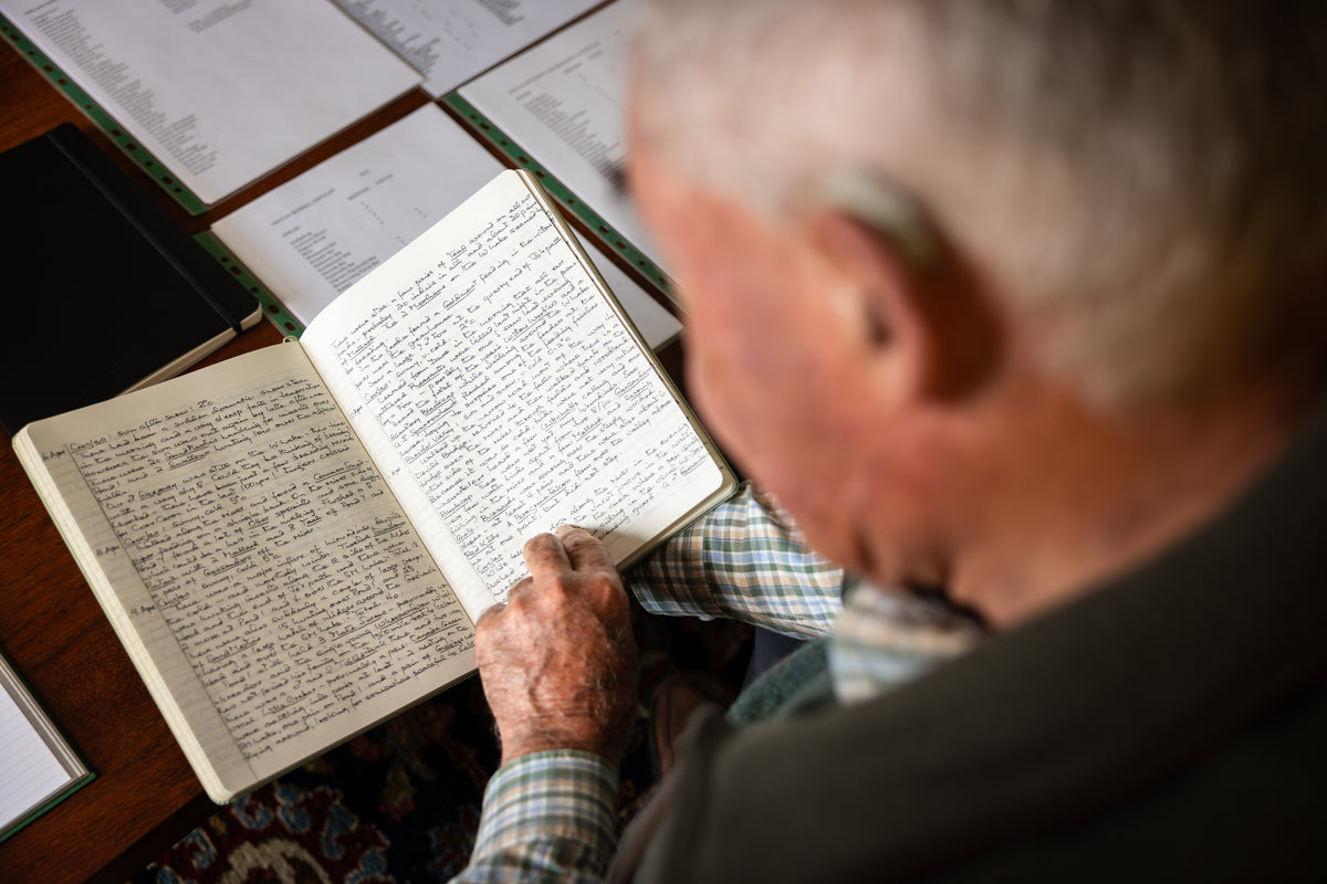 Elderly man reading a handwritten notebook at a wooden table with papers.