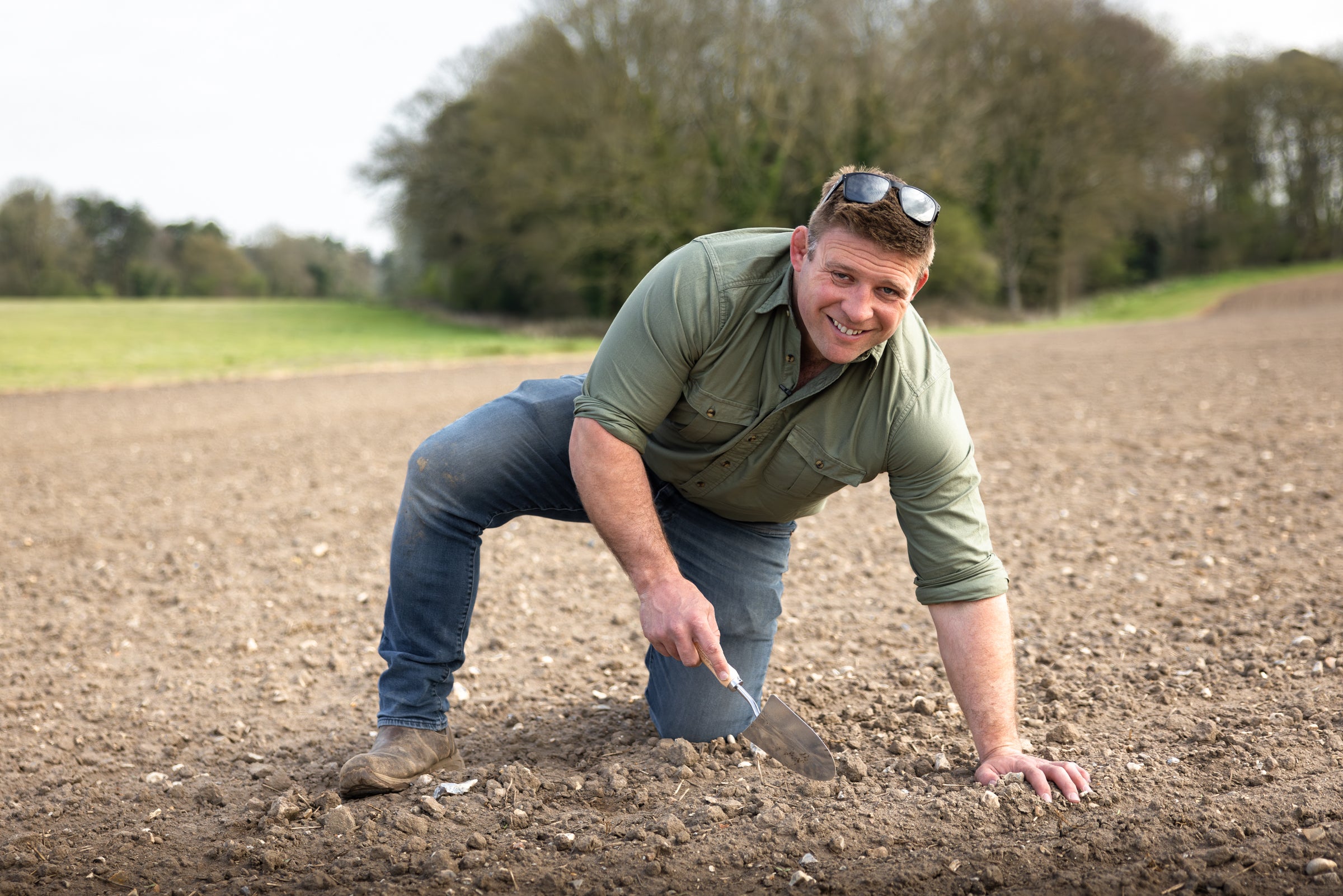 Man in a green shirt and jeans crouching in a plowed field with trees in the background.