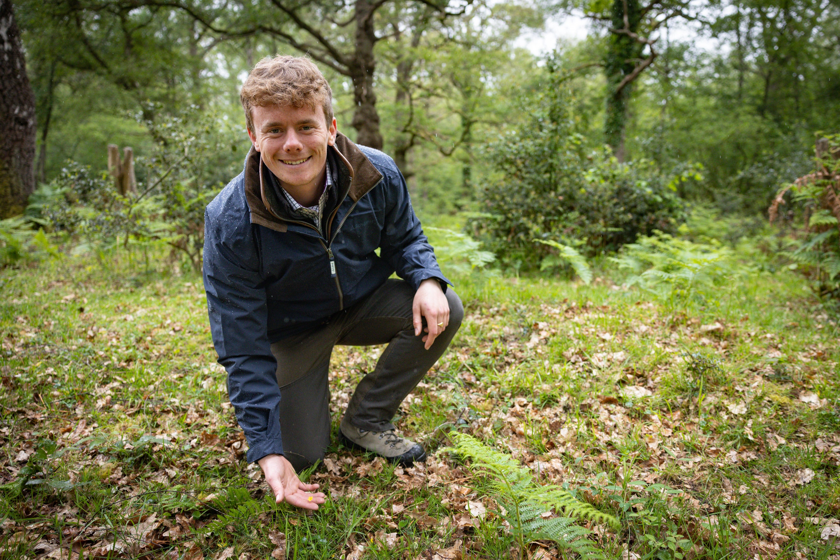 Tom Hilder crouching in a wooded outdoor setting, wearing a navy blue zip-up jacket, dark trousers, and hiking boots, with his hand reaching toward green plants on the ground.