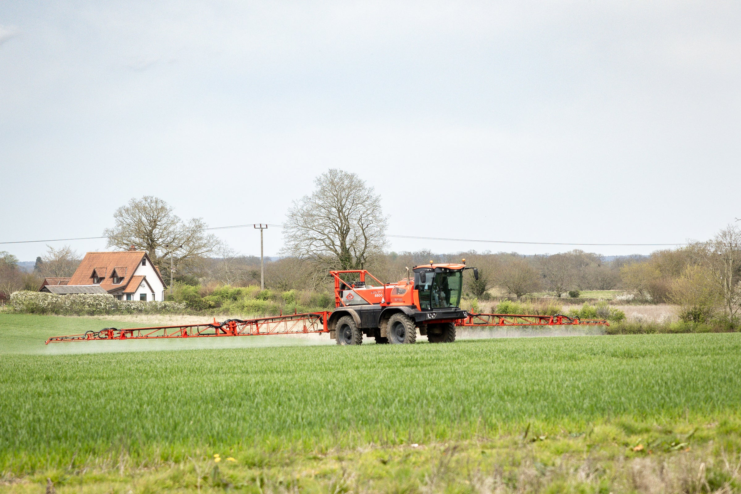 Red agricultural sprayer in a field with a house and trees in the background