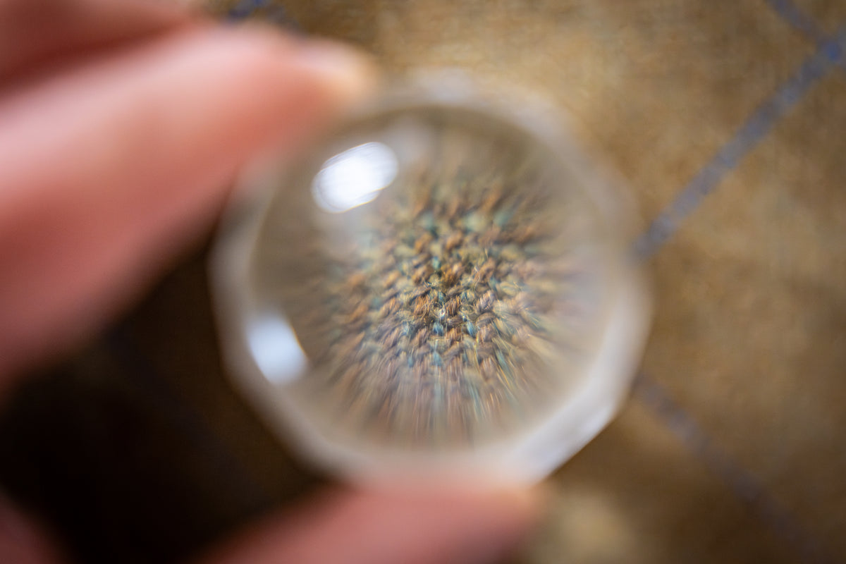 A close-up of a hand holding a  magnifying glass over tweed fabric