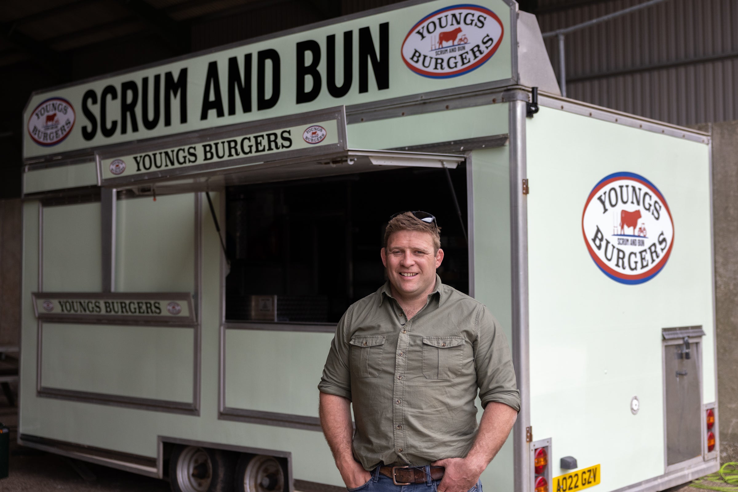 Man standing in front of a food truck with 'Scrum and Bun' and 'Youngs Burgers' branding.
