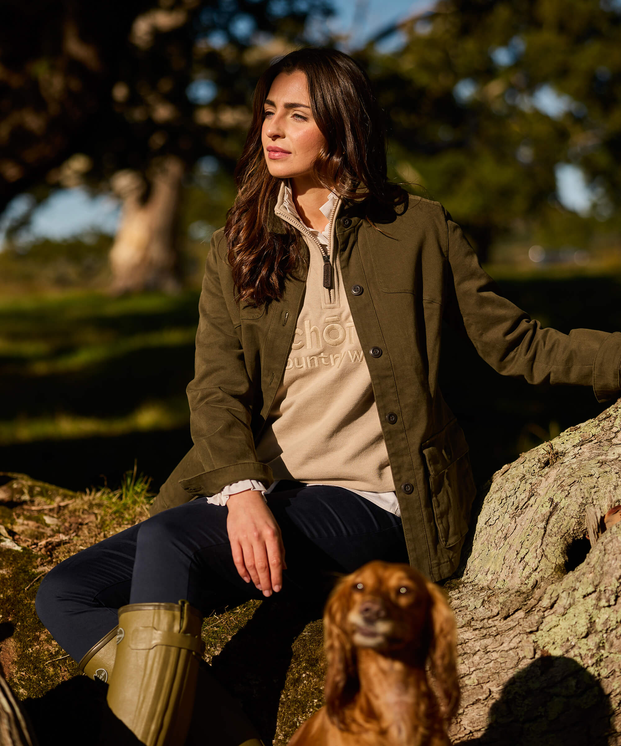 Woman wearing an olive green stretch cotton utility jacket over a cream Schöffel quarter-zip sweatshirt and navy trousers, seated on a mossy rock in a sunlit parkland setting with a red cocker spaniel in the foreground.