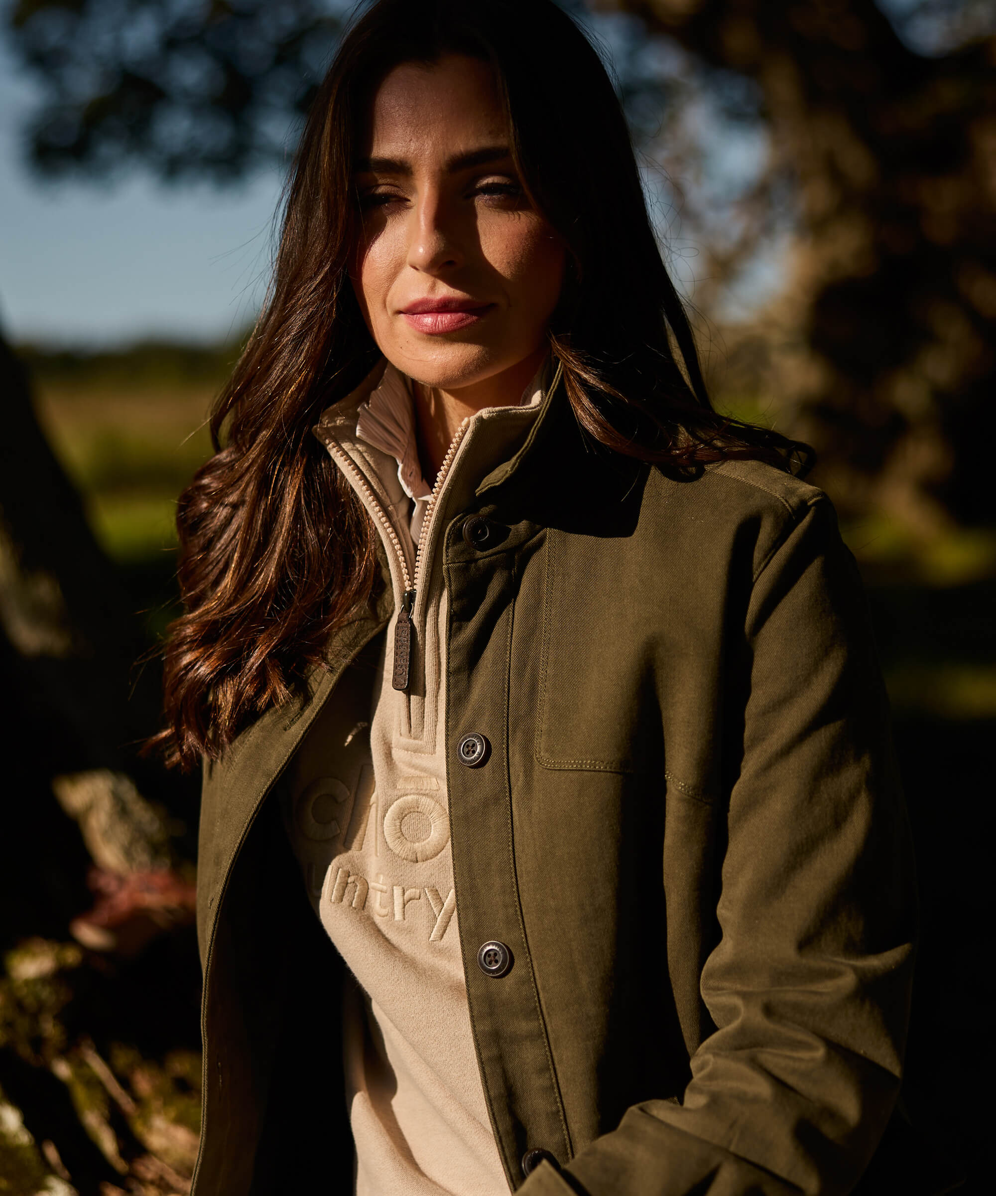 Close-up of a woman wearing an olive green button-front utility jacket with a chest pocket and horn buttons, layered over a cream Schöffel sweatshirt, photographed in warm autumn light against a woodland backdrop.