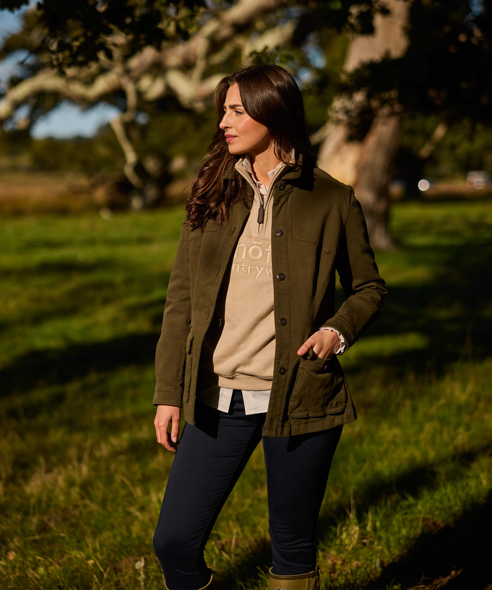 Woman in an olive green stretch cotton utility jacket with large front pockets and an adjustable waist, styled with navy trousers and khaki Wellington boots, standing in a sunlit meadow beneath a large oak tree.