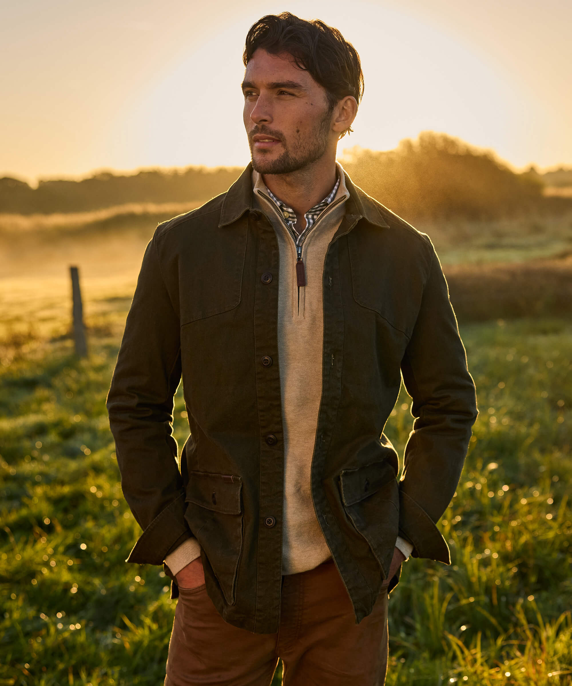 Man wearing an olive green cotton utility jacket with chest pockets and large flap front pockets, layered over a cream quarter-zip knitwear and gingham shirt with rust-coloured chinos, standing in a misty field at golden hour.