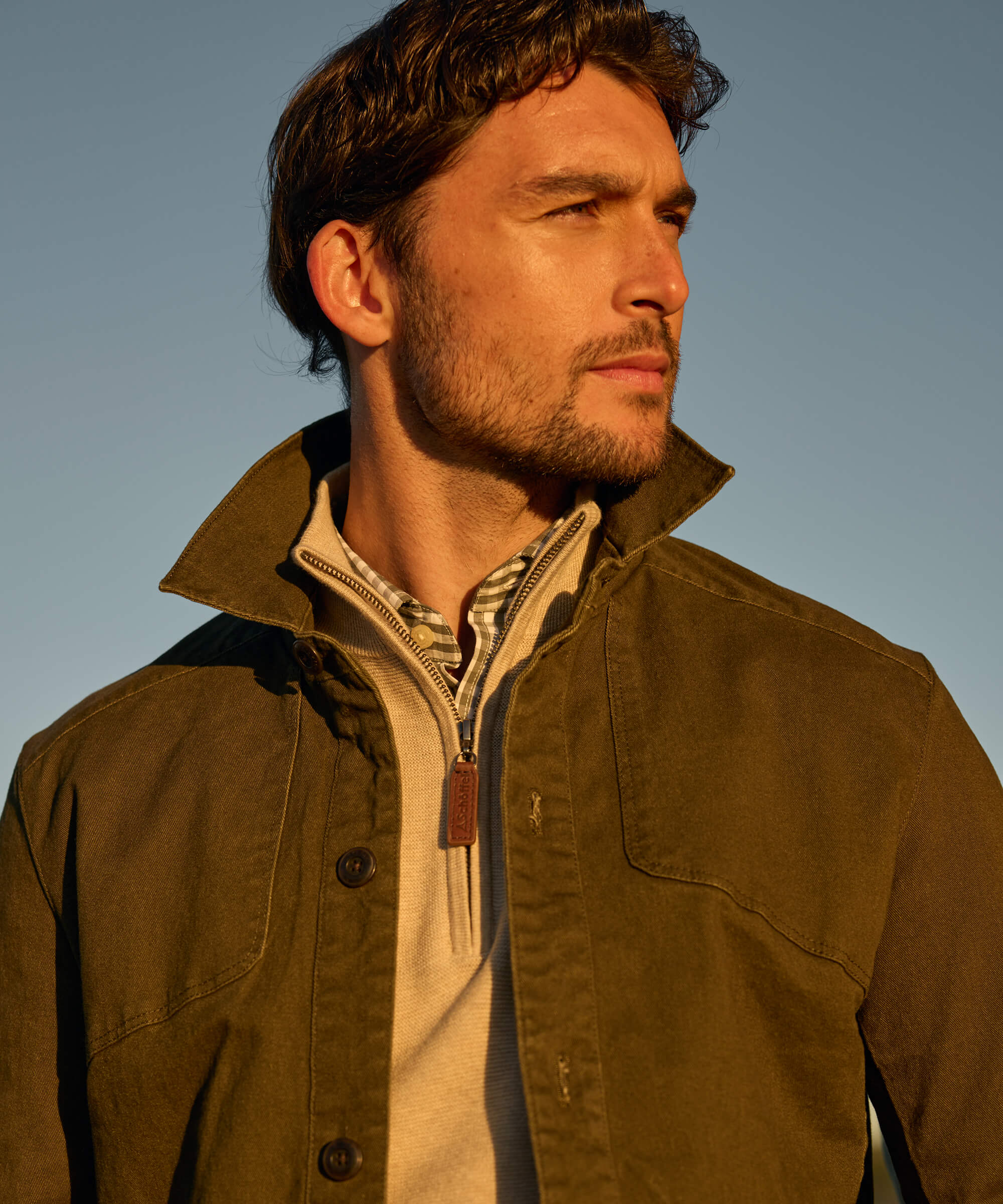 Close-up portrait of a man wearing an olive green utility jacket with a structured collar and horn buttons, layered over a cream quarter-zip and gingham shirt, photographed against a pale blue sky in warm late-afternoon light