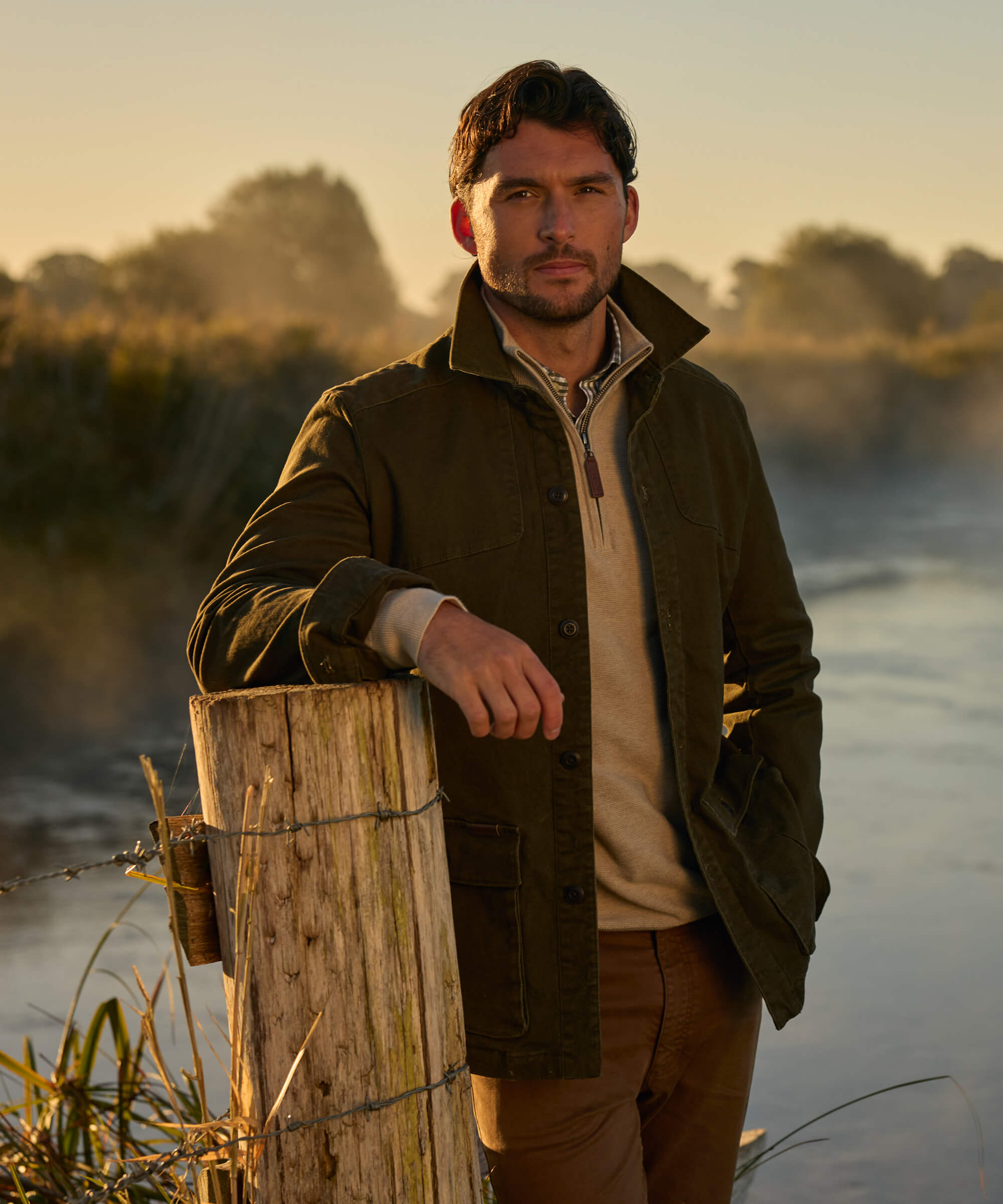 Man in an olive green cotton utility jacket leaning on a weathered wooden fence post beside a misty river at sunrise, styled with a cream quarter-zip and rust-coloured chinos.