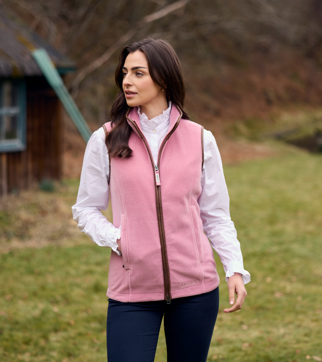 Person wearing a pink fleece gilet with zip fastening over a white long‑sleeve shirt, standing outdoors on grass.