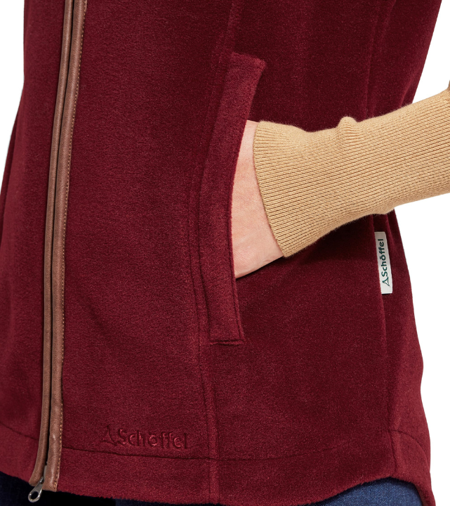 Close-up detail of a woman's hand in the side pocket of a claret red fleece gilet with tan leather-trim zip and embroidered Schöffel logo at the hem, worn over a mustard yellow knitted jumper.