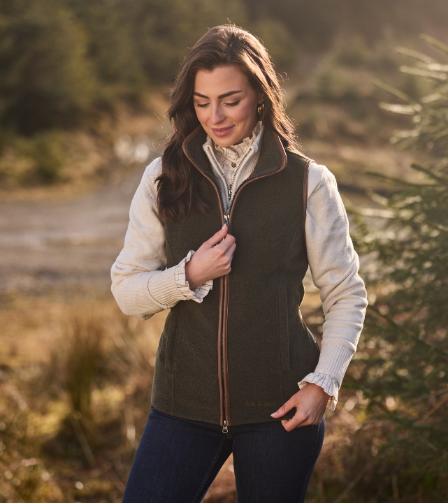 Woman wearing a dark green fleece gilet over a cream shirt, standing outdoors in a countryside setting.