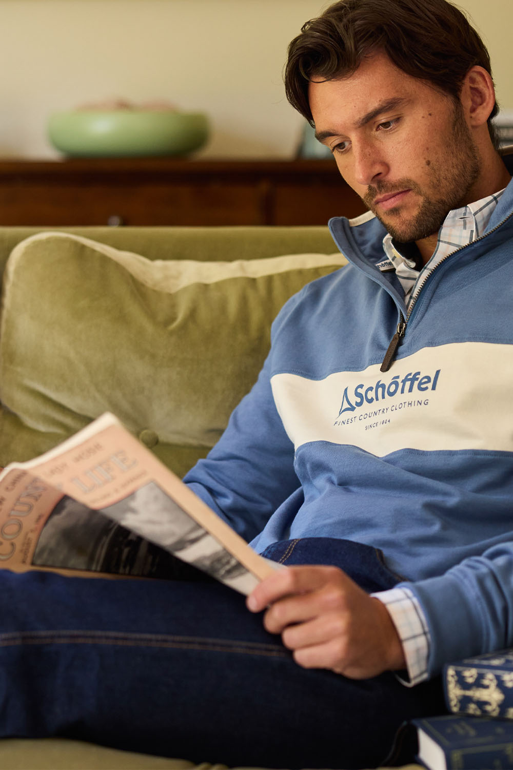Man wearing a blue jacket with 'Schöffel' logo, reading a newspaper on a couch.