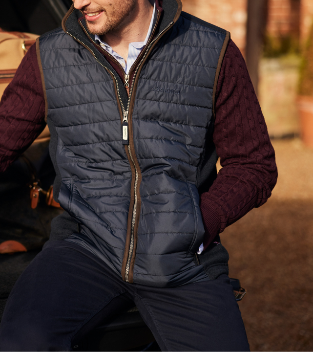 A man seated outdoors wearing a petrol blue quilted gilet with brown leather-effect zip trim and a tonal Schöffel logo embroidered on the chest. He is layered over a burgundy cable-knit jumper and a blue check shirt, paired with navy trousers.