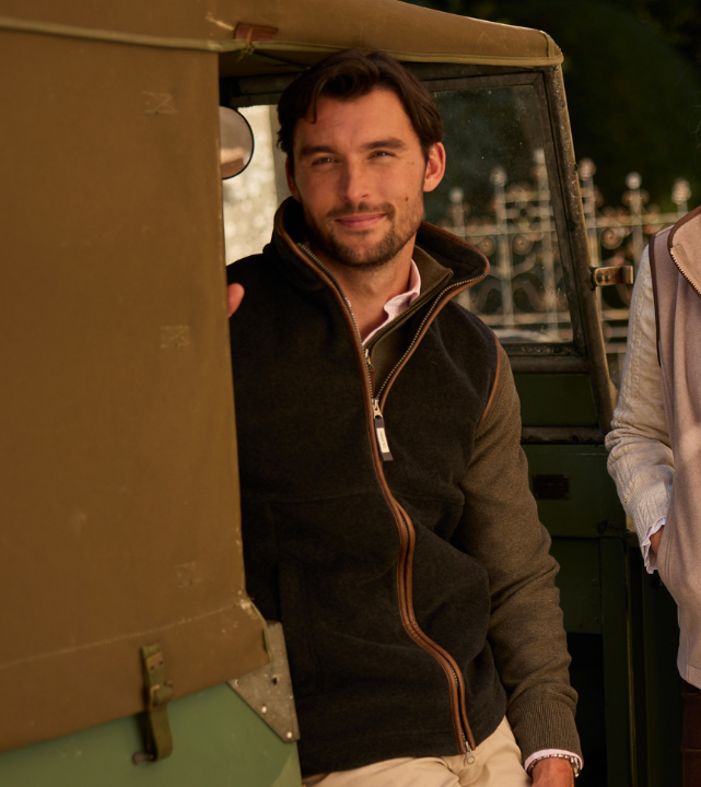 Person wearing a dark olive fleece gilet with zip fastening, standing outdoors beside a vehicle in a countryside setting.