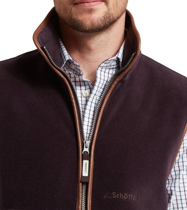 Close-up of a man wearing a port red fleece gilet with tan leather-trim full-length zip and embroidered Schöffel logo on the chest, worn open over a pastel checked shirt.