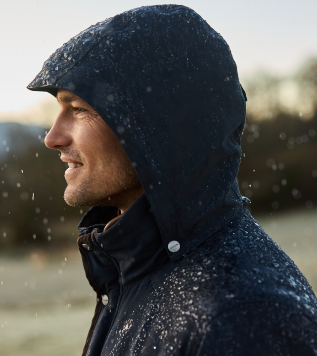 Profile view of a person wearing a carbon navy hooded country coat during rainfall outdoors.